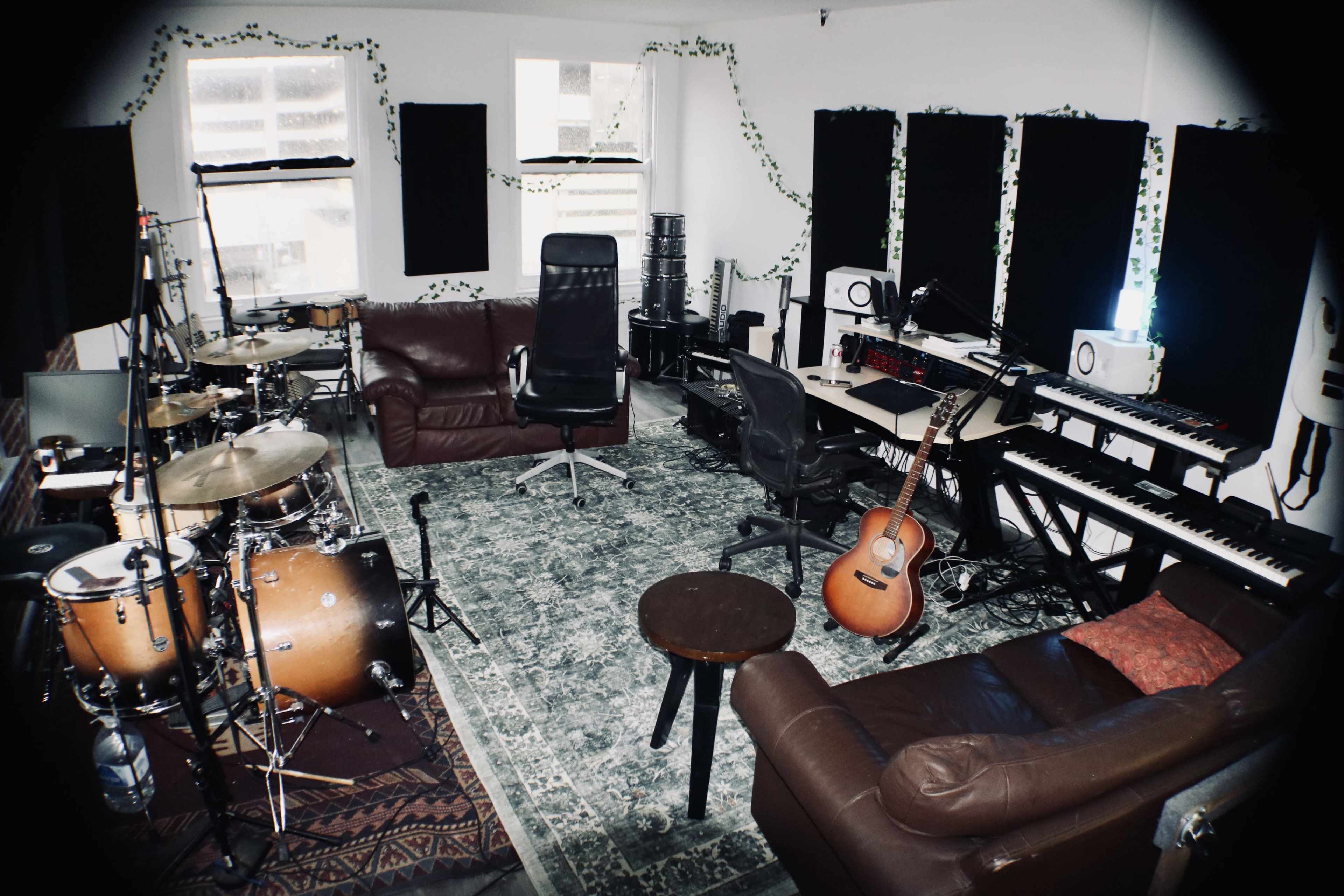 The image shows a music studio with a drum set, keyboard, guitar, and seating arranged around a patterned rug.