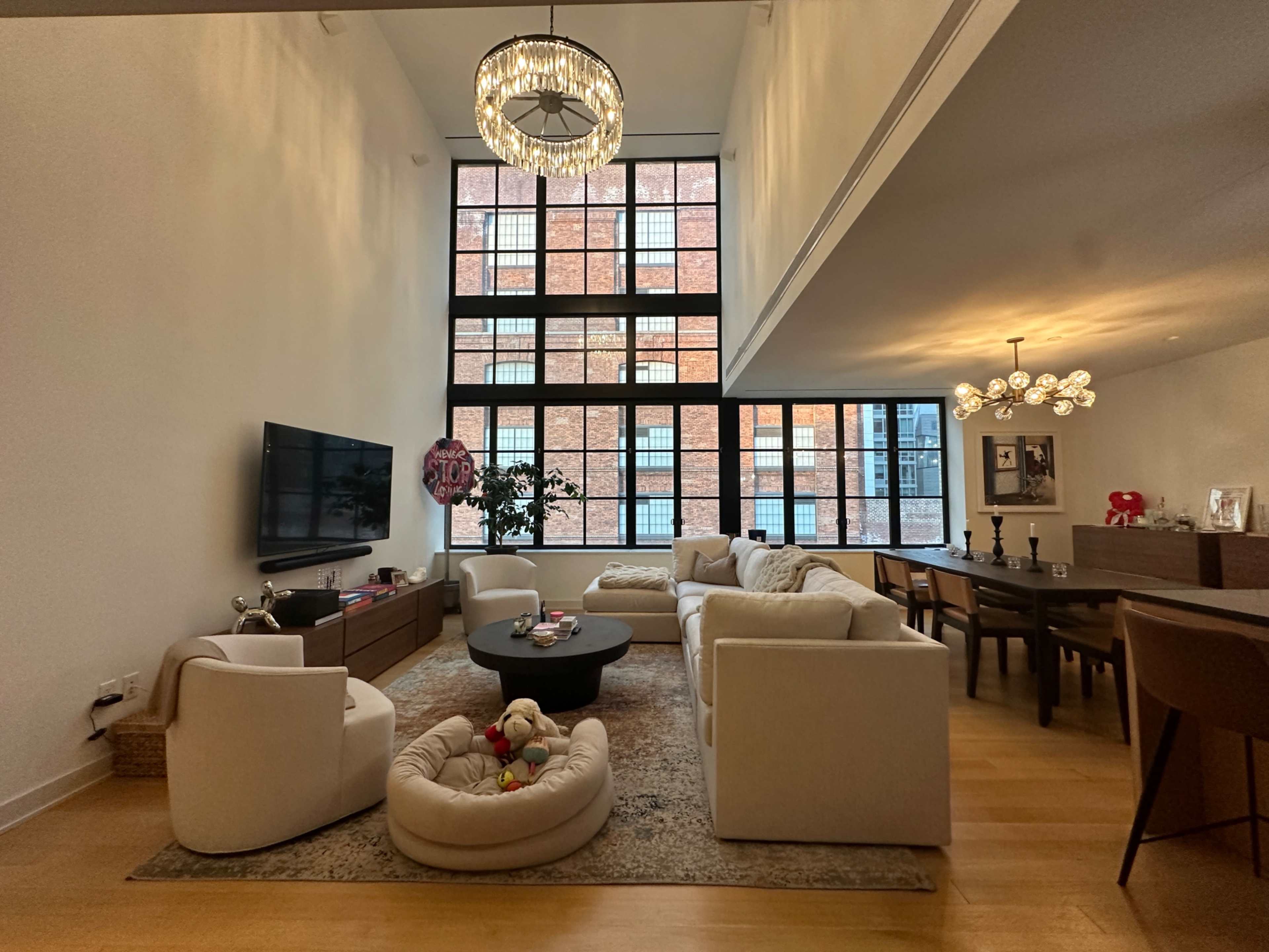 The image shows a spacious living area with large windows, a modern seating arrangement, and a dining table, featuring a decorative chandelier and a pet bed on the floor.