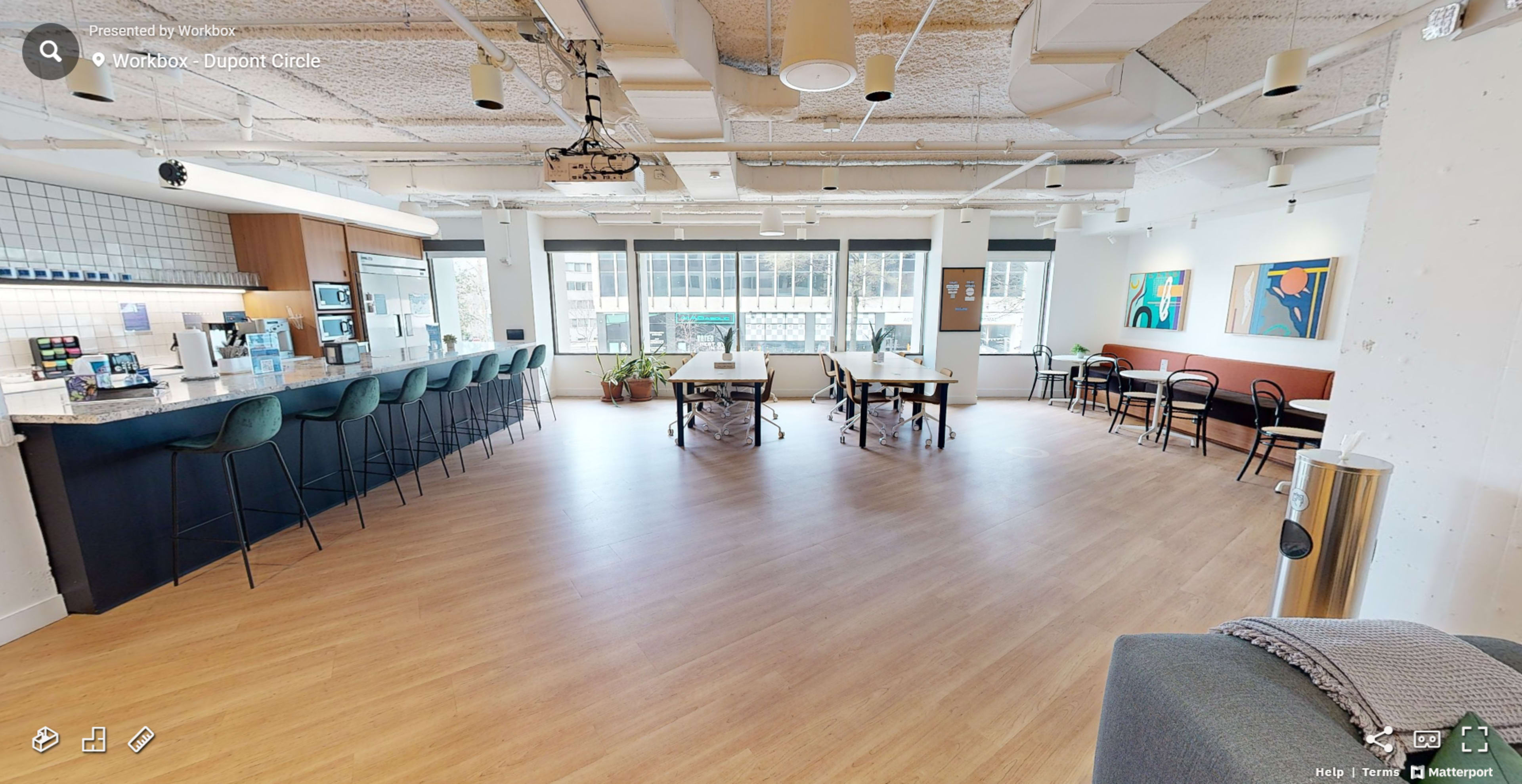 The image shows a modern, open workspace with a kitchen area, two long tables, and seating, all bathed in natural light from large windows.