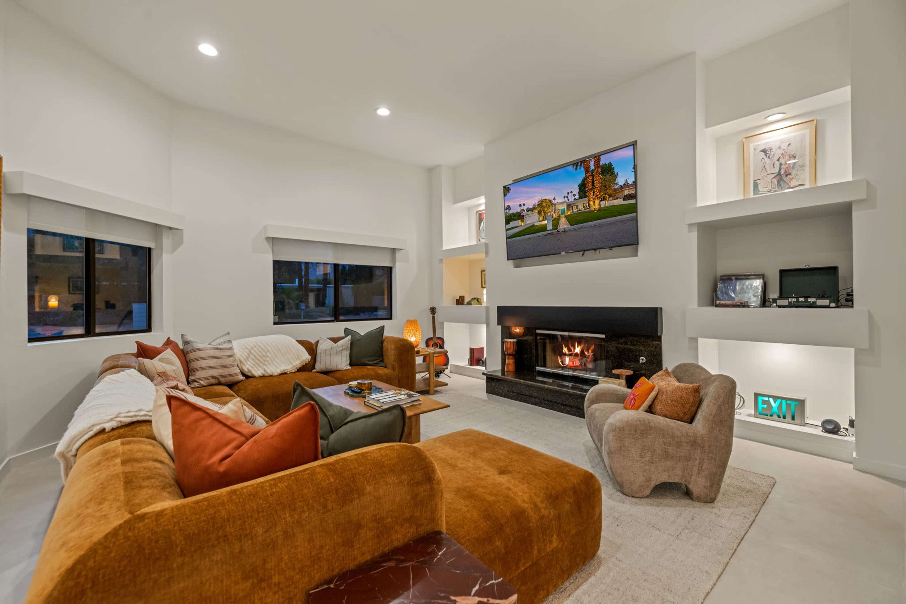 The living room features a large sectional sofa, a flat-screen TV mounted on the wall, and a fireplace, all arranged in a bright, modern space.