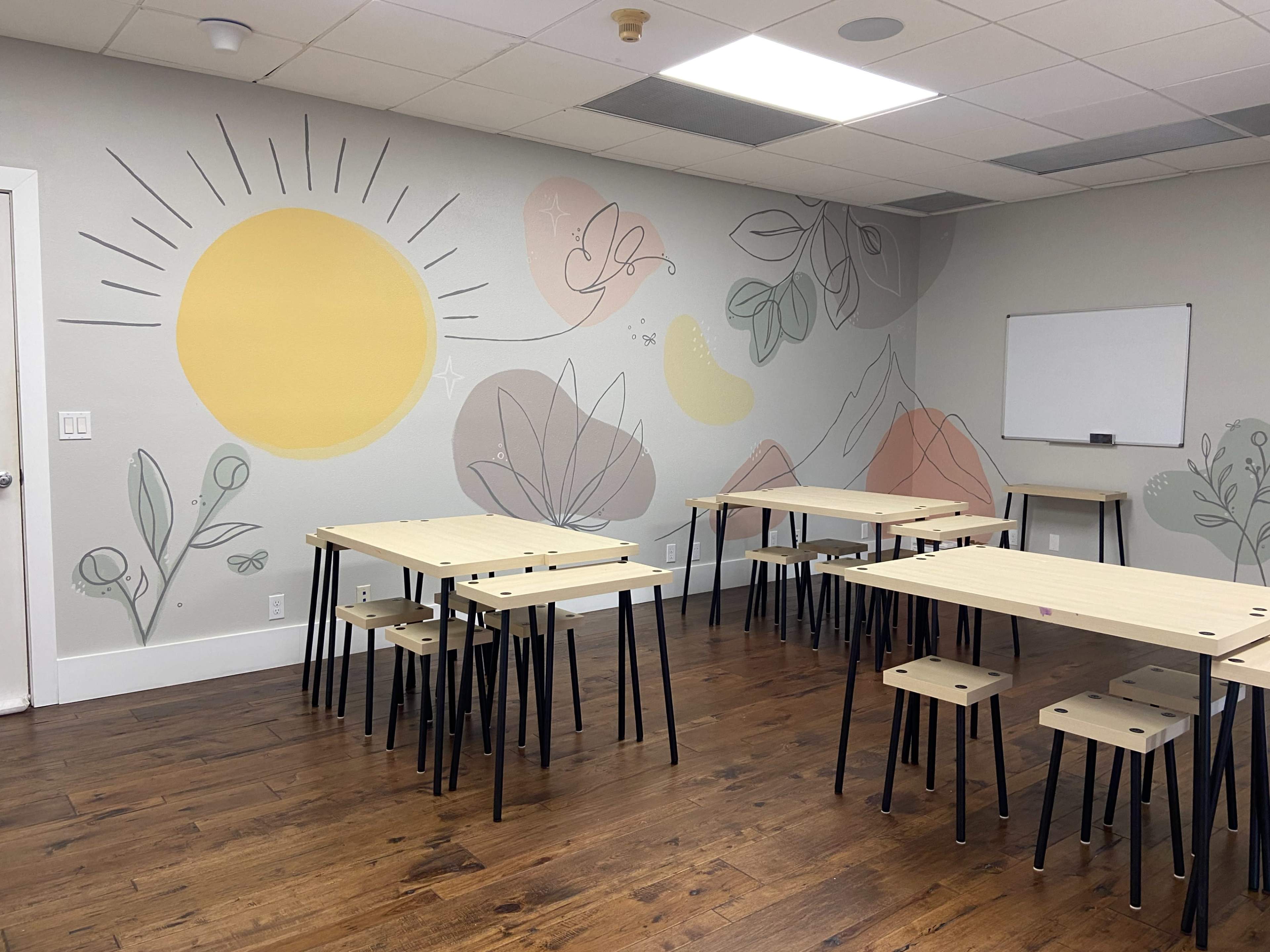 The room features light-colored walls with a mural of a sun and abstract plants, along with several wooden tables and stools arranged for seating.