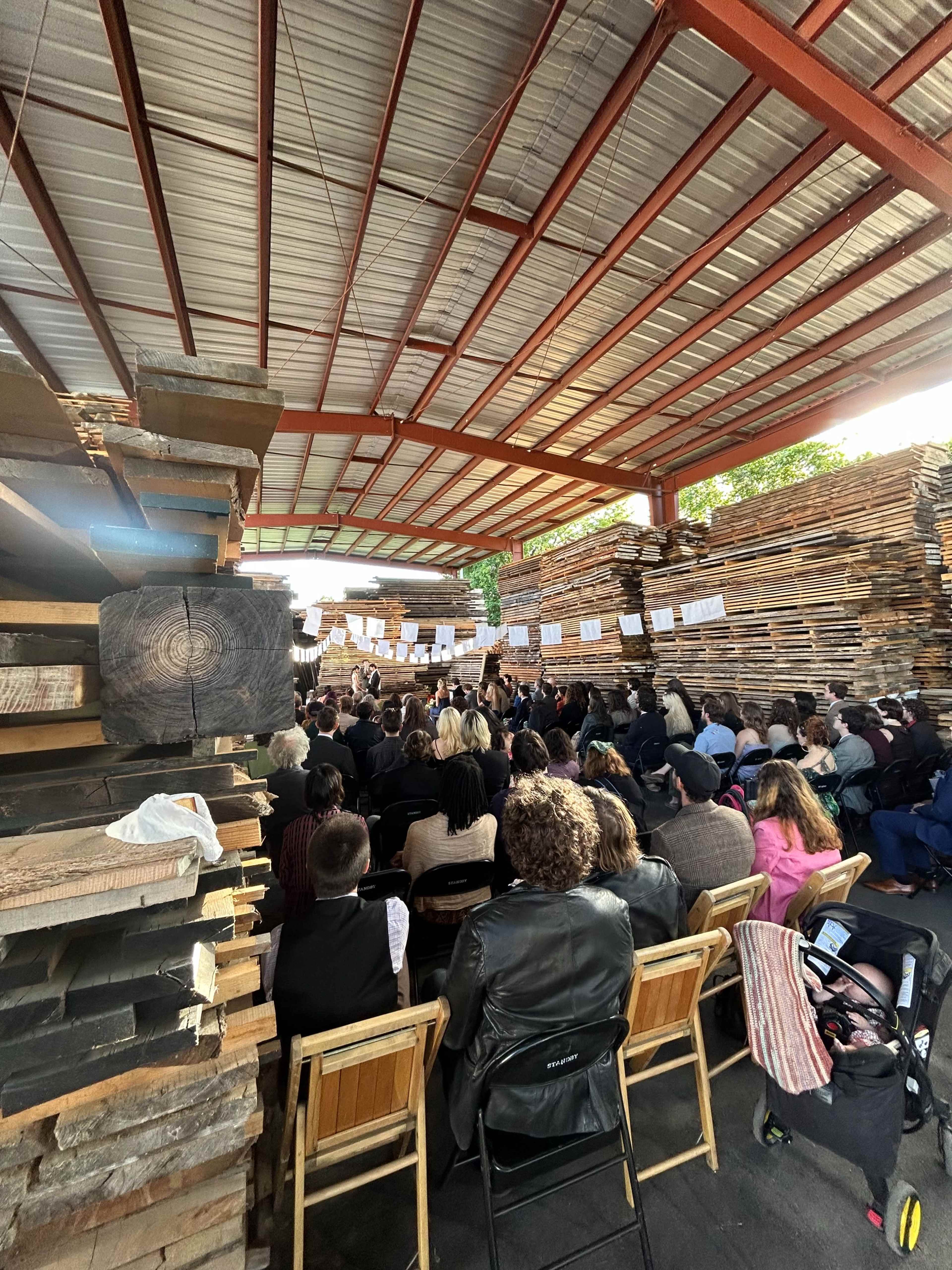 A large audience sits in a warehouse filled with stacked wooden planks, attending a presentation or performance.