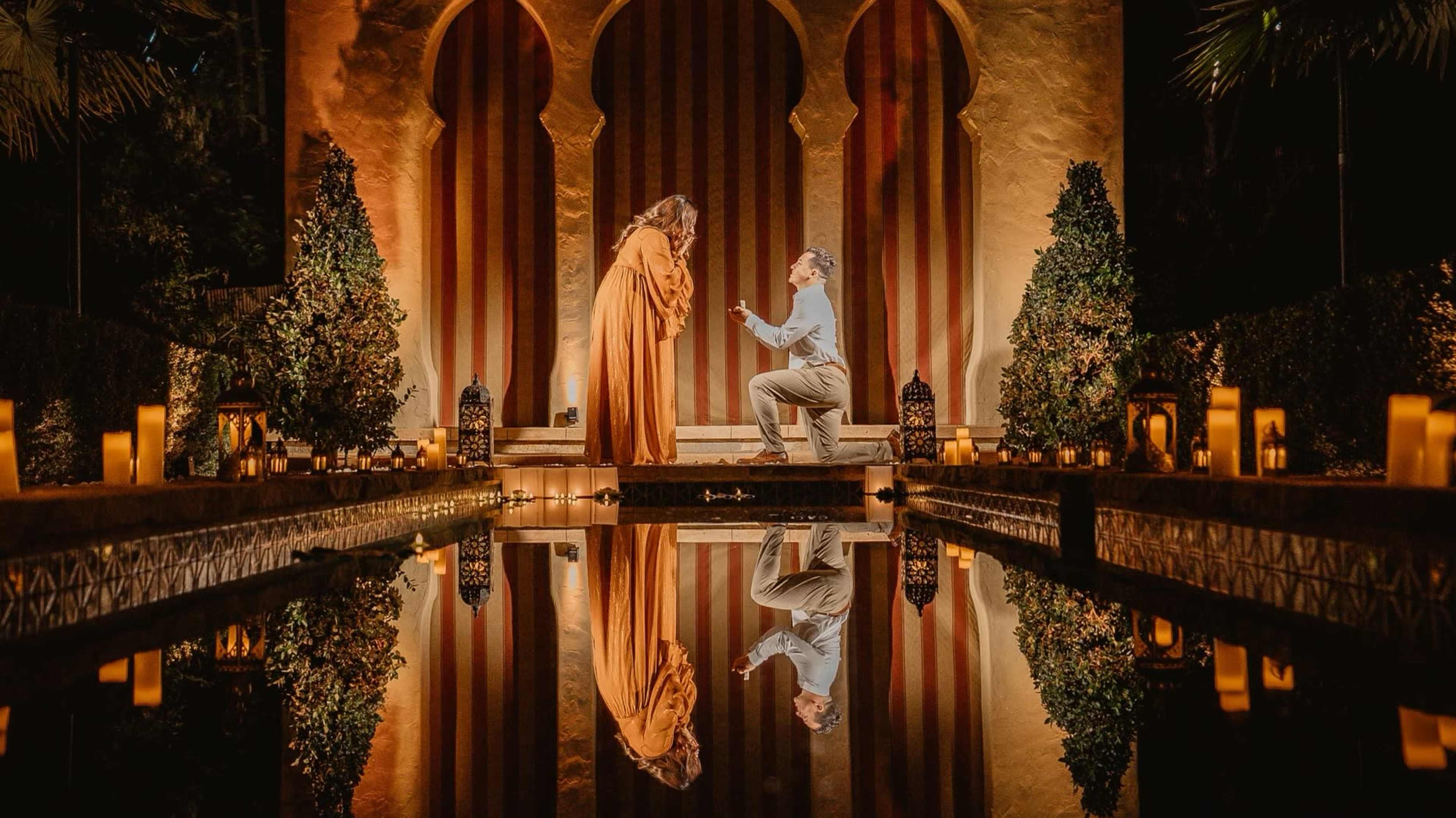 A man is proposing to a woman in an ornate garden setting, surrounded by lanterns and reflecting in a water feature.