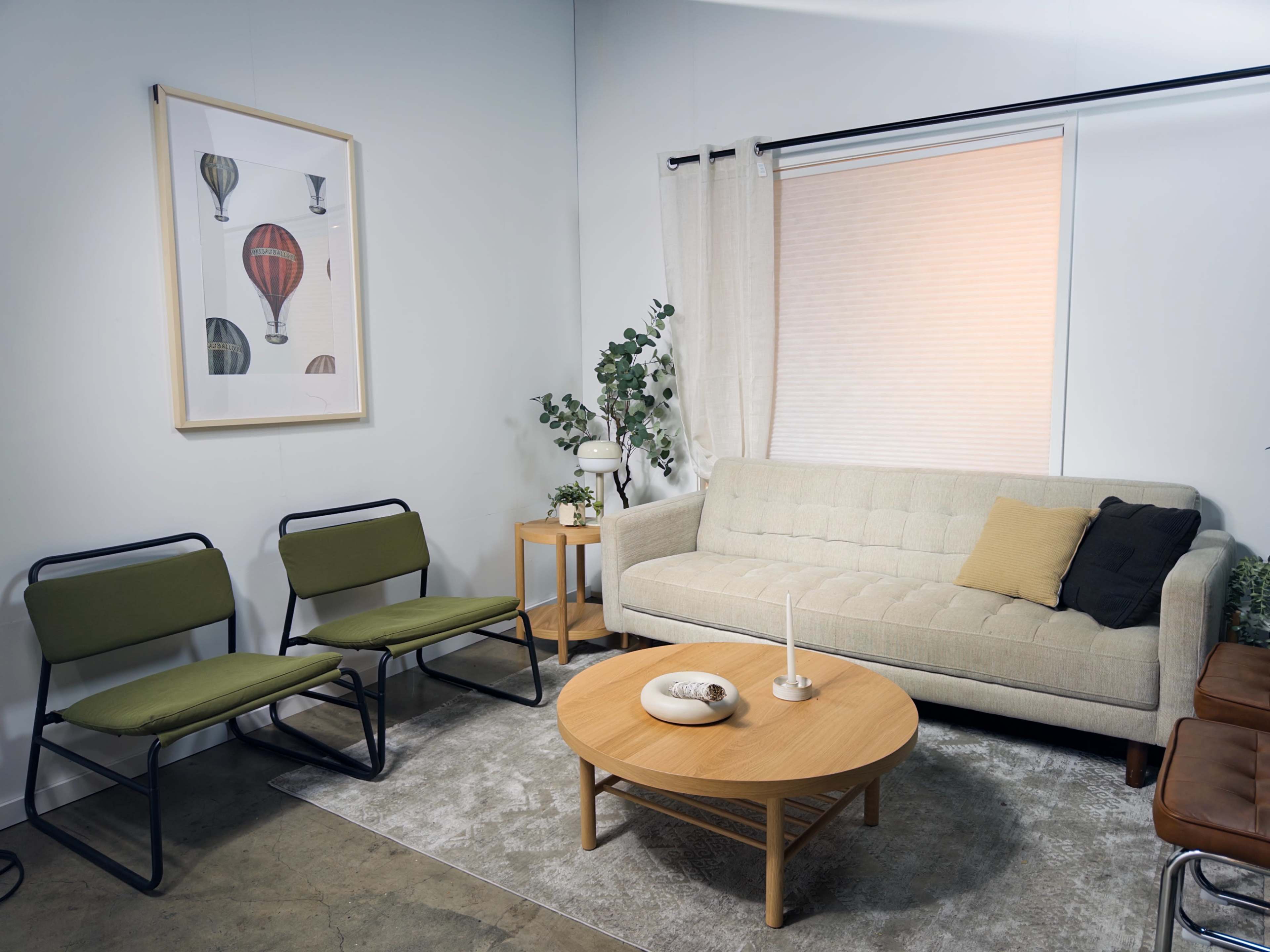 The image shows a neatly arranged living room with a beige couch, two green chairs, a round wooden coffee table, and a framed illustration of hot air balloons on the wall.