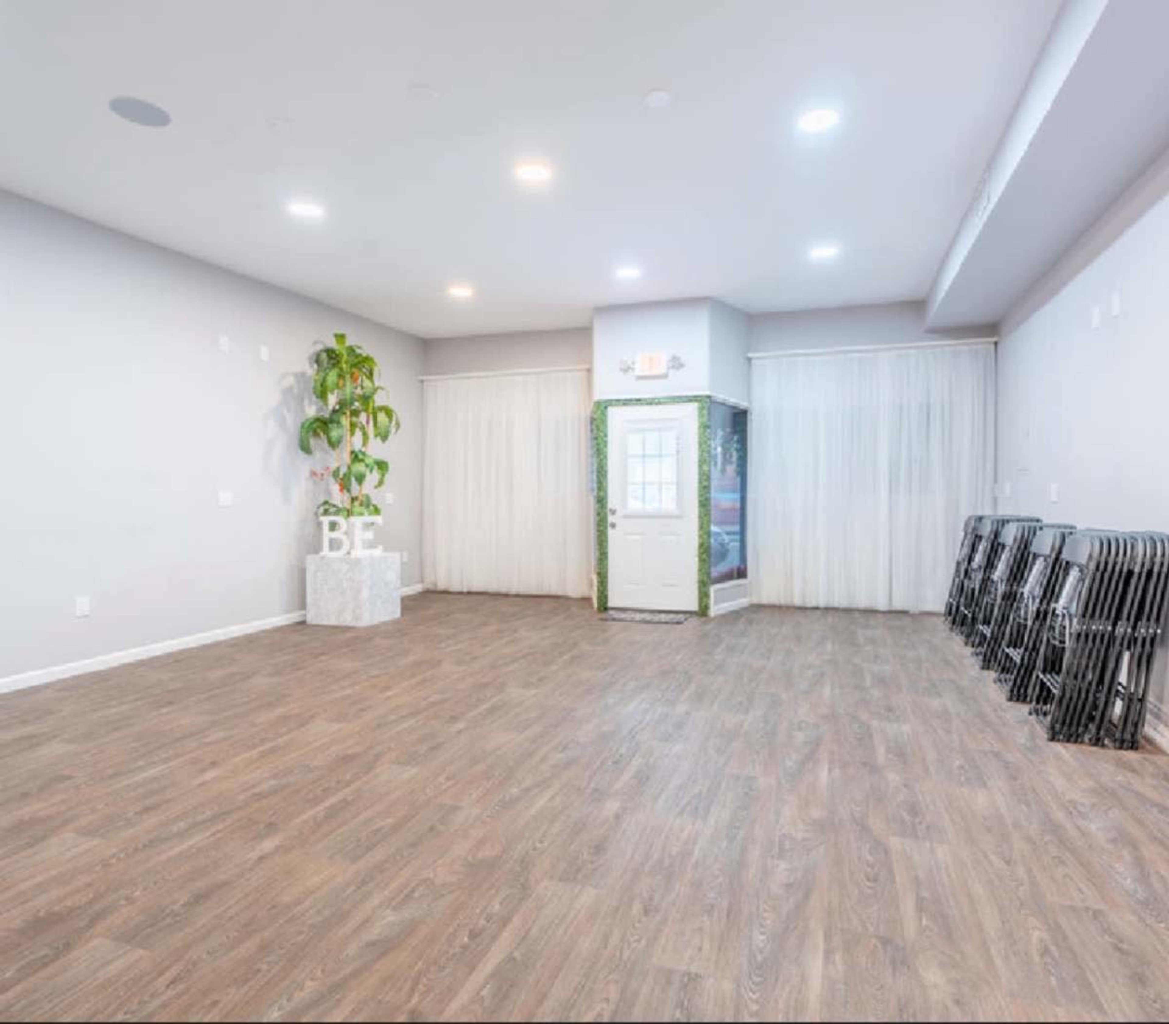 An empty room with wooden flooring, a potted plant in the corner, a door with curtains, and several folding chairs stacked against a wall.