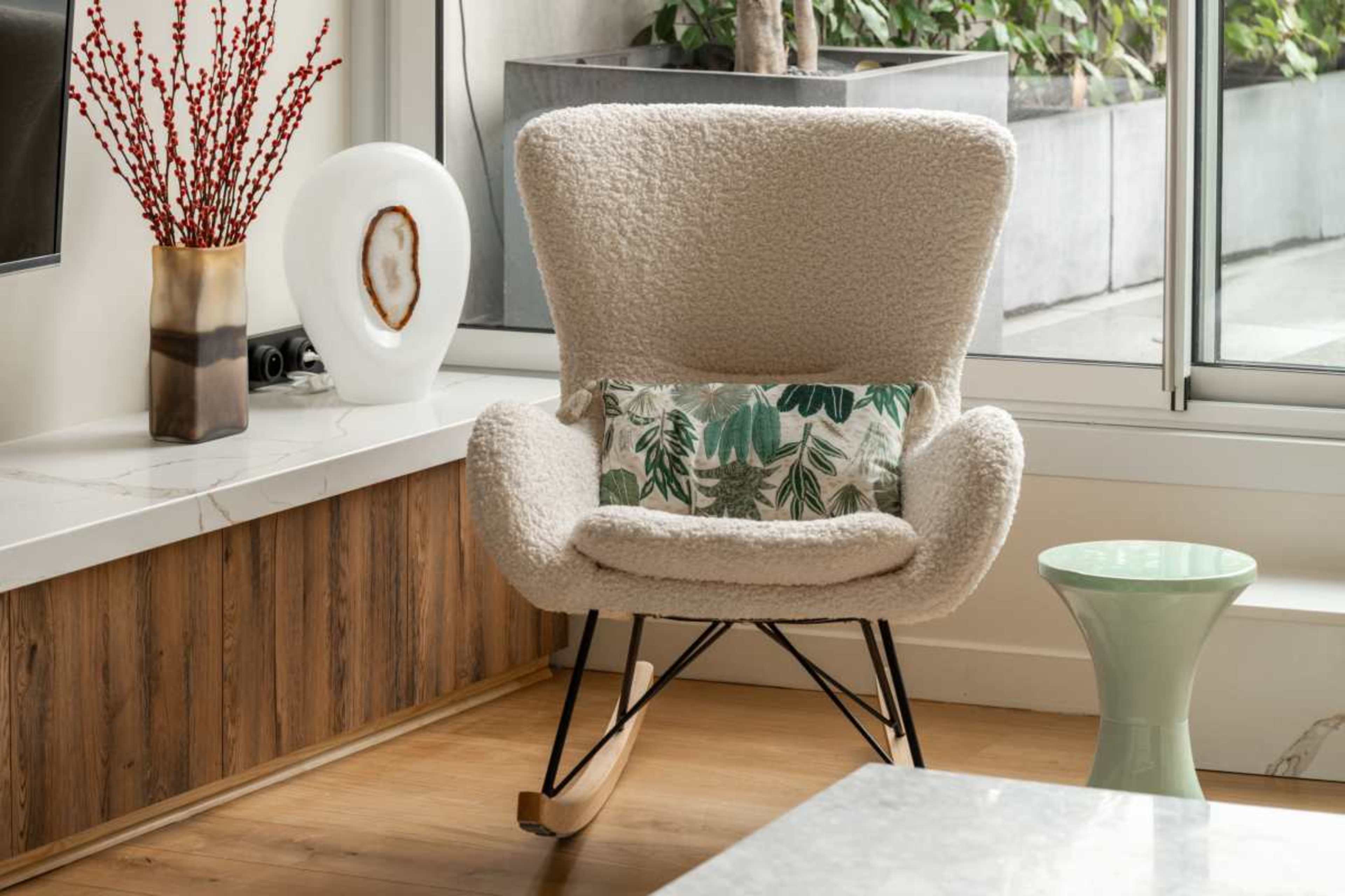 The image features a cozy, light-colored rocking chair with a decorative pillow, positioned next to a small green side table and surrounded by a modern interior setting.