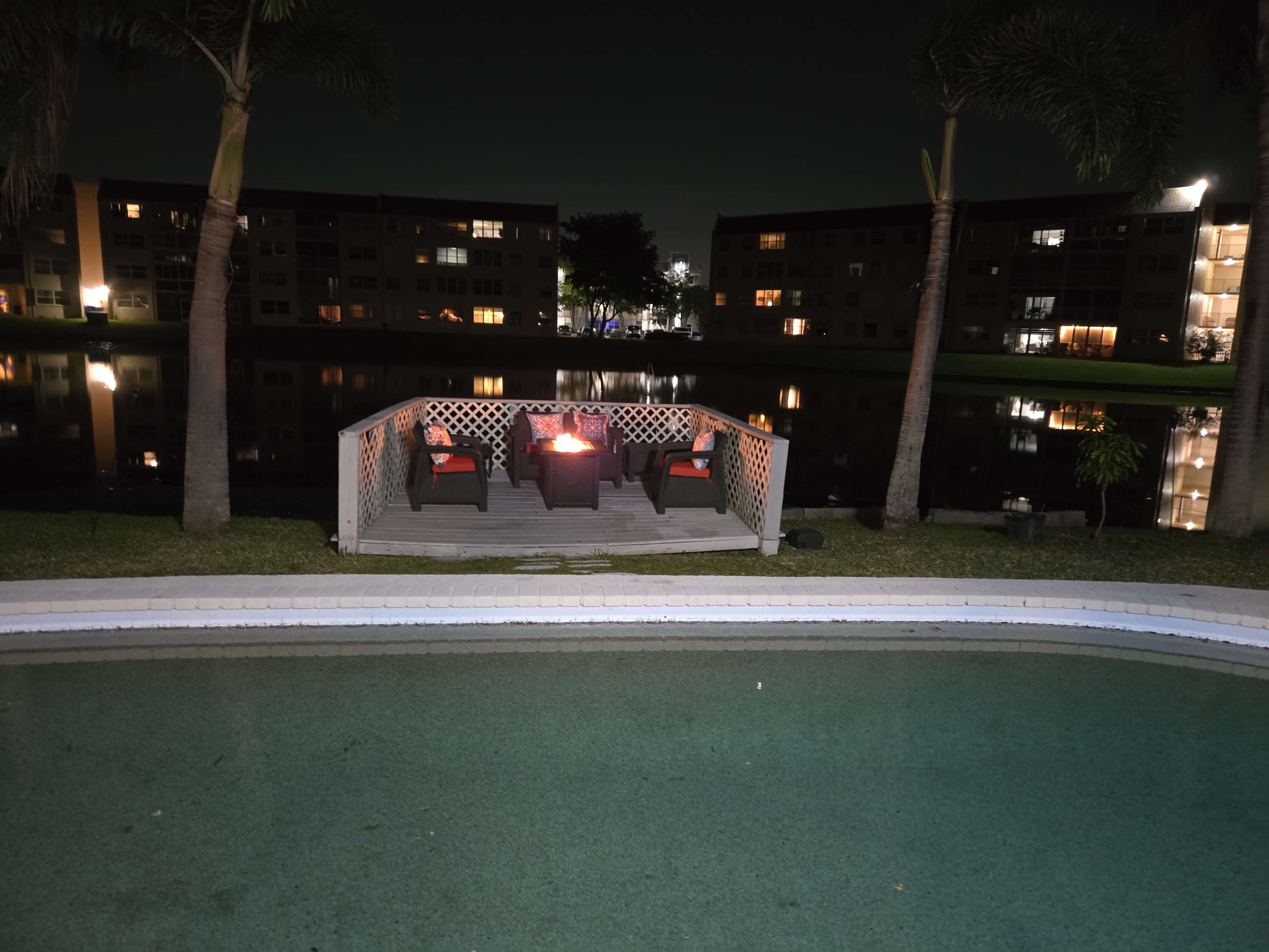 The image shows a wooden deck with four chairs and a fire pit, situated by a calm body of water reflecting nearby apartment buildings at night.
