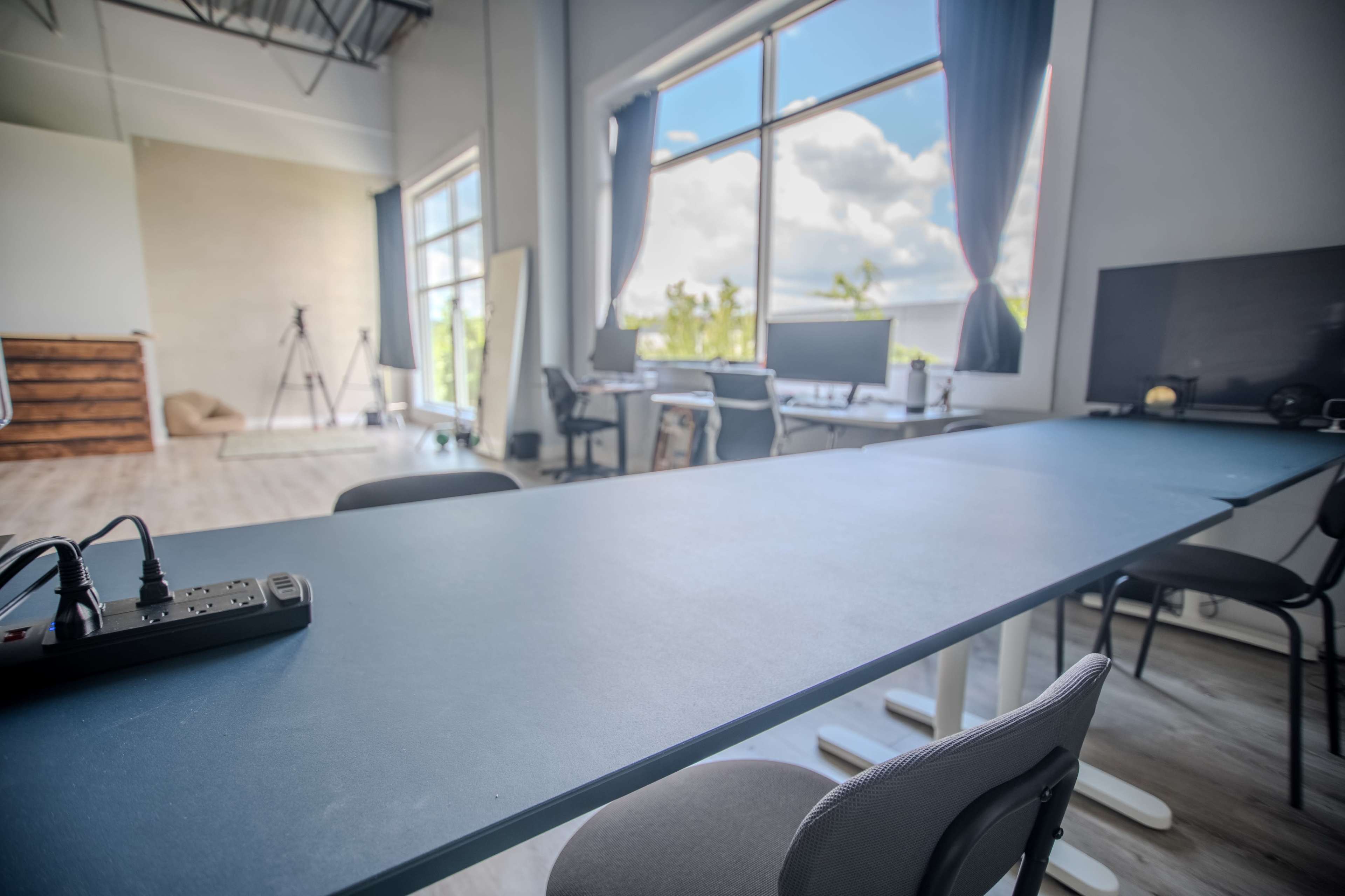 The image shows a modern workspace with a long desk, chairs, and large windows allowing natural light to enter.
