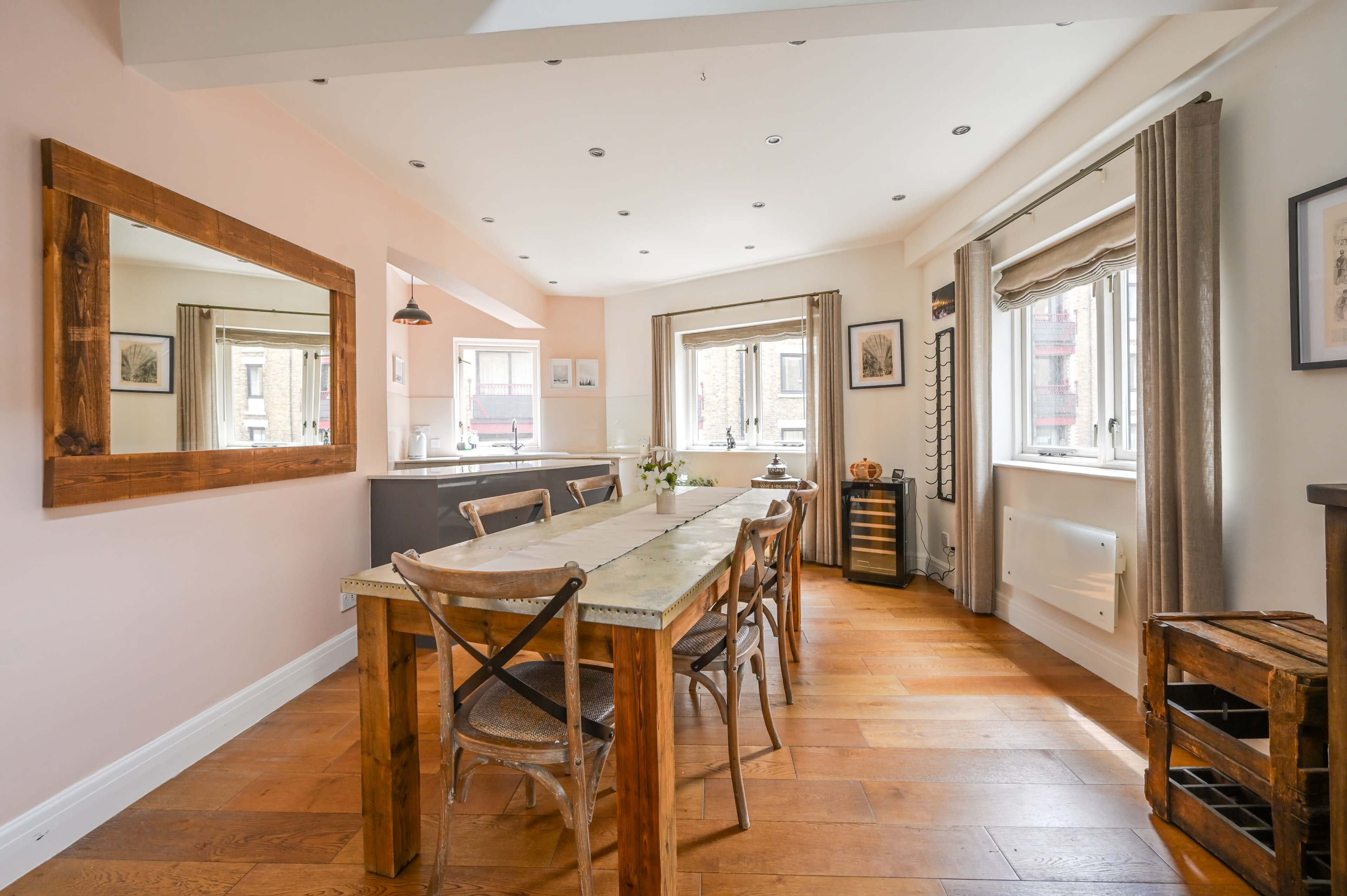 The image shows a modern kitchen and dining area with a wooden table, chairs, and large windows providing natural light.
