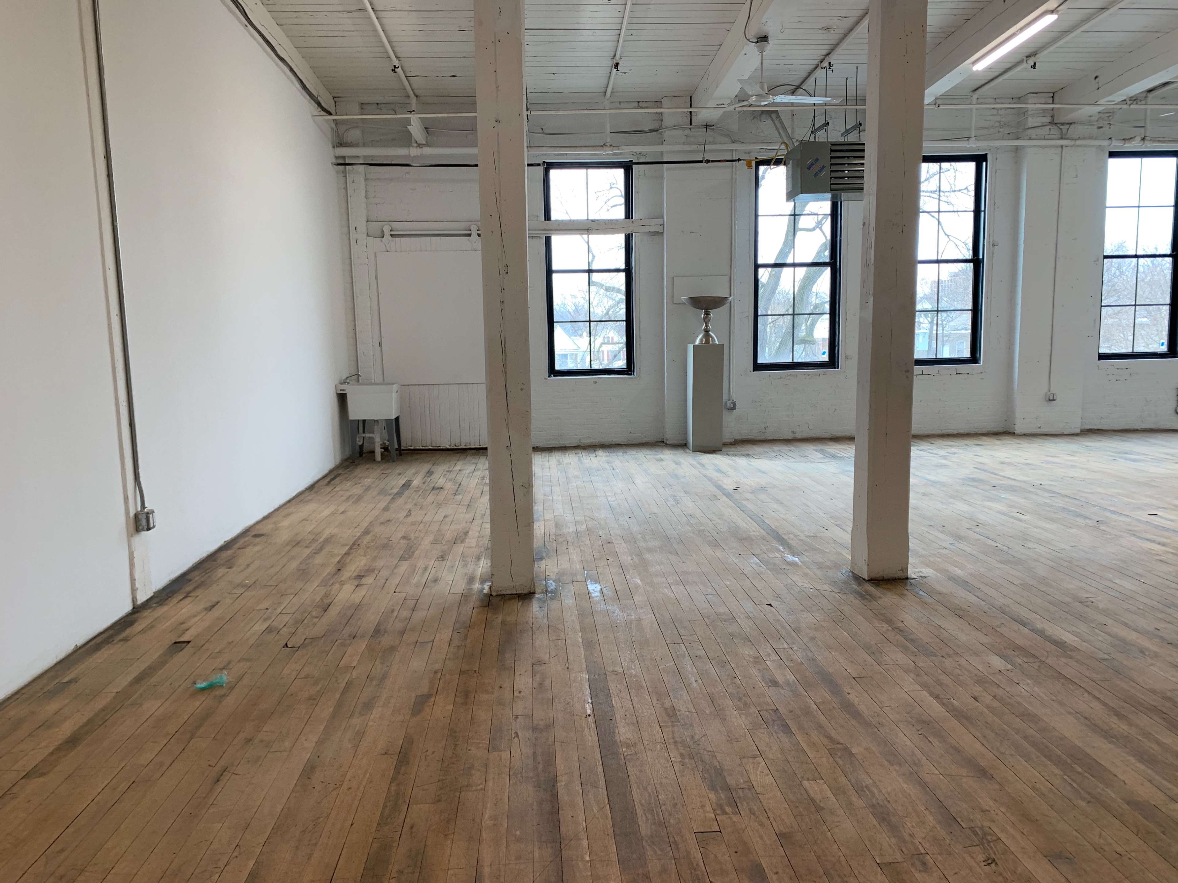 An empty room with wooden flooring, white walls, large windows, and exposed beams.