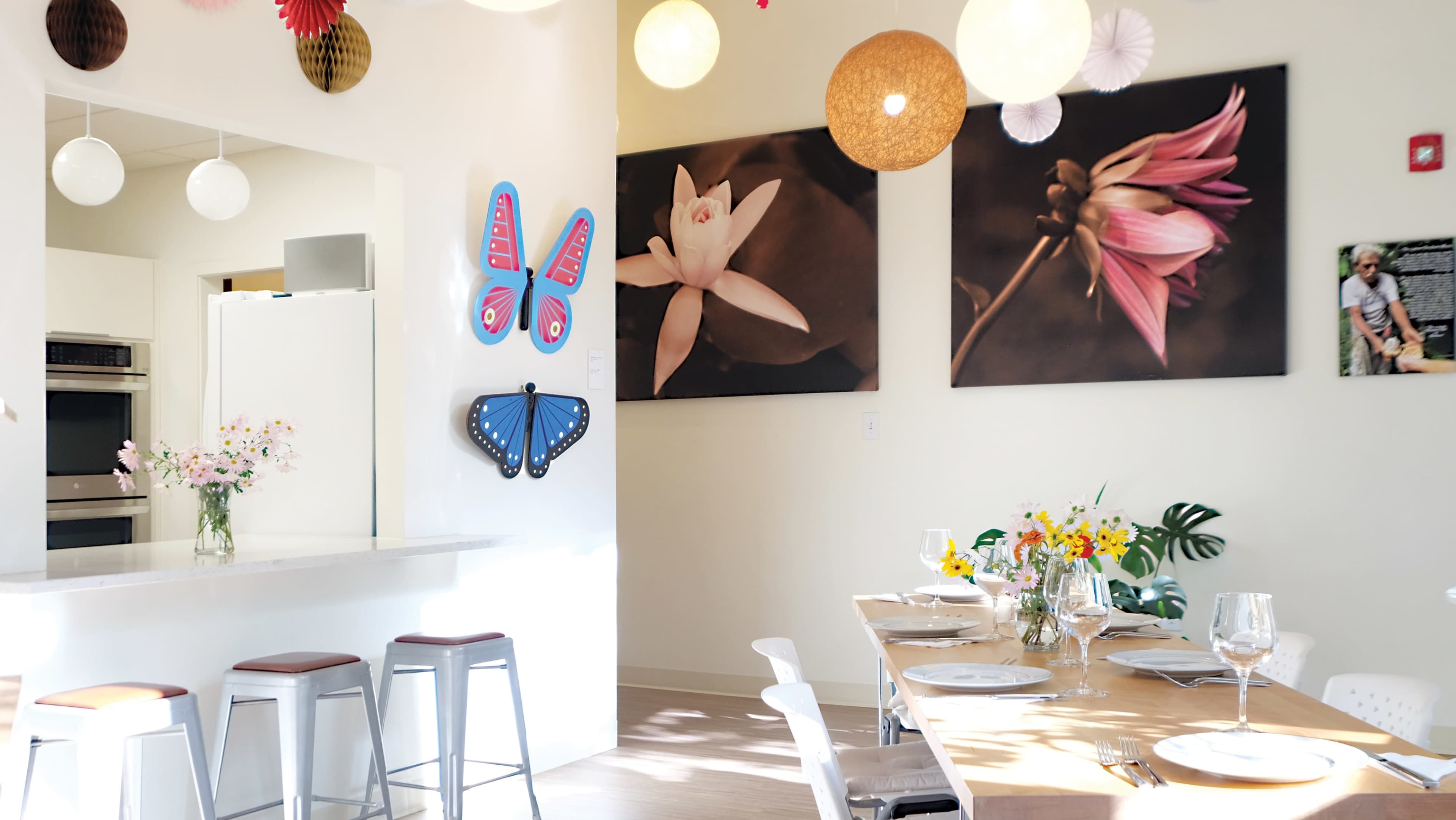 A bright dining area with a wooden table set for a meal, surrounded by colorful artwork, flowers, and various hanging light fixtures.