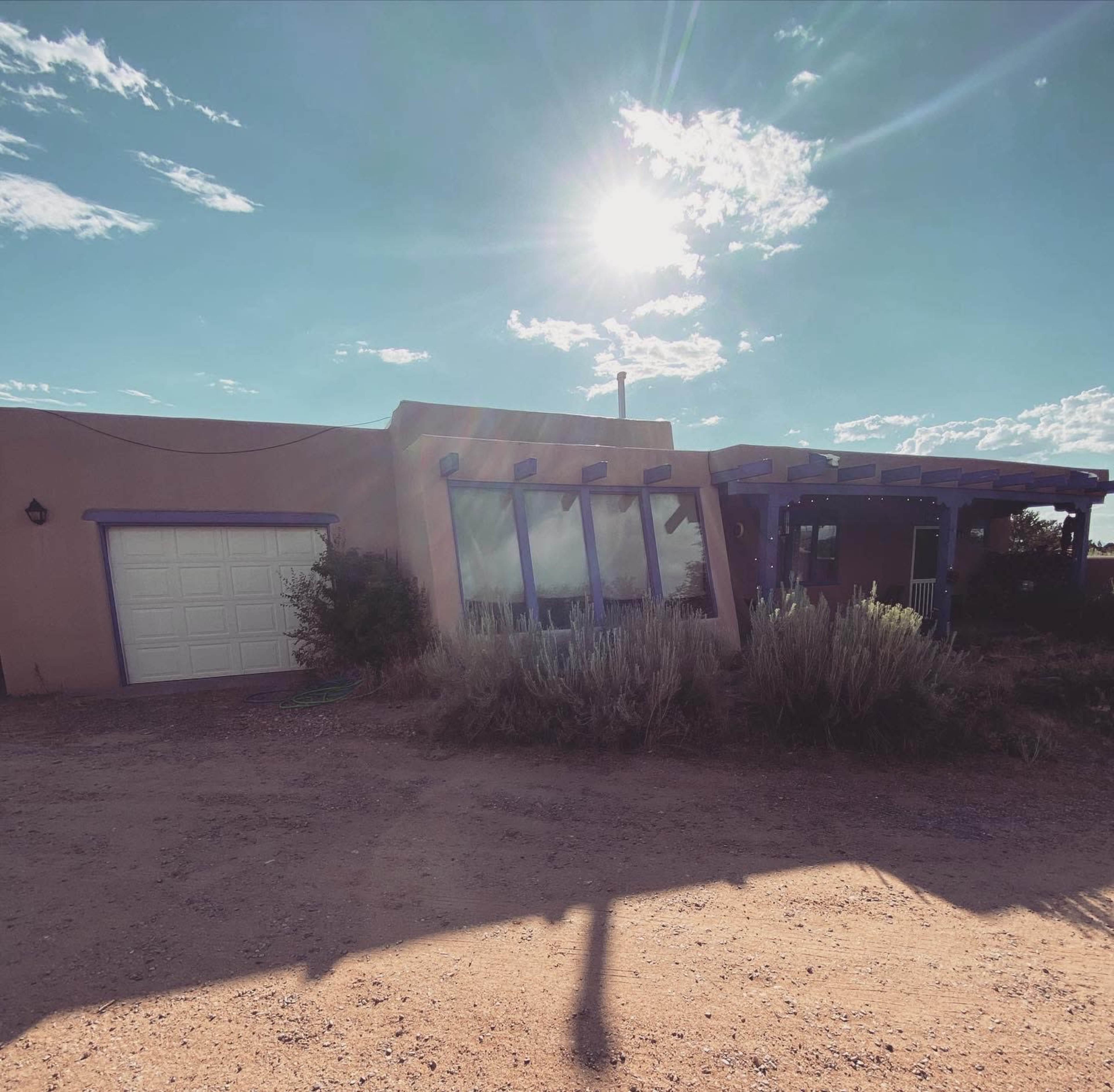 A single-story adobe house with a large window and a garage door is nestled among desert vegetation under a bright sun.