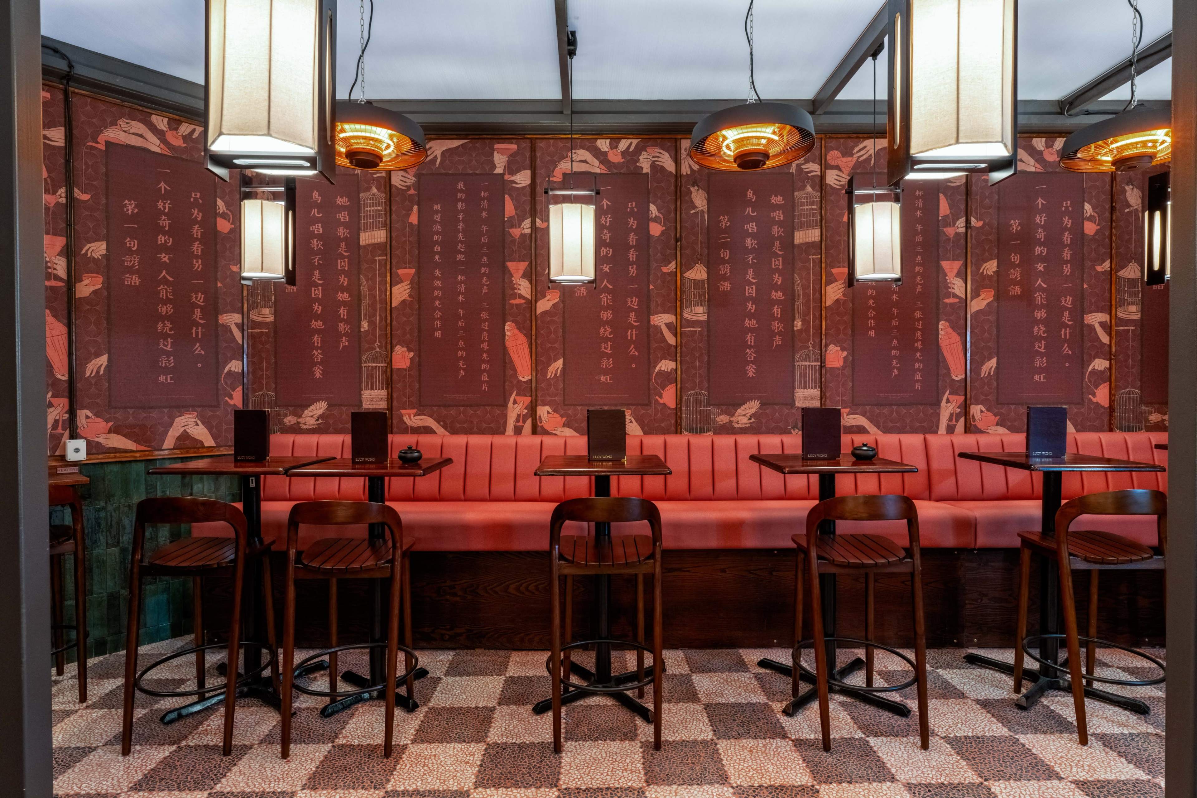 The image shows a modern restaurant interior featuring red upholstered seating along the walls, wooden tables and chairs, and decorative wall art with text in a traditional script.