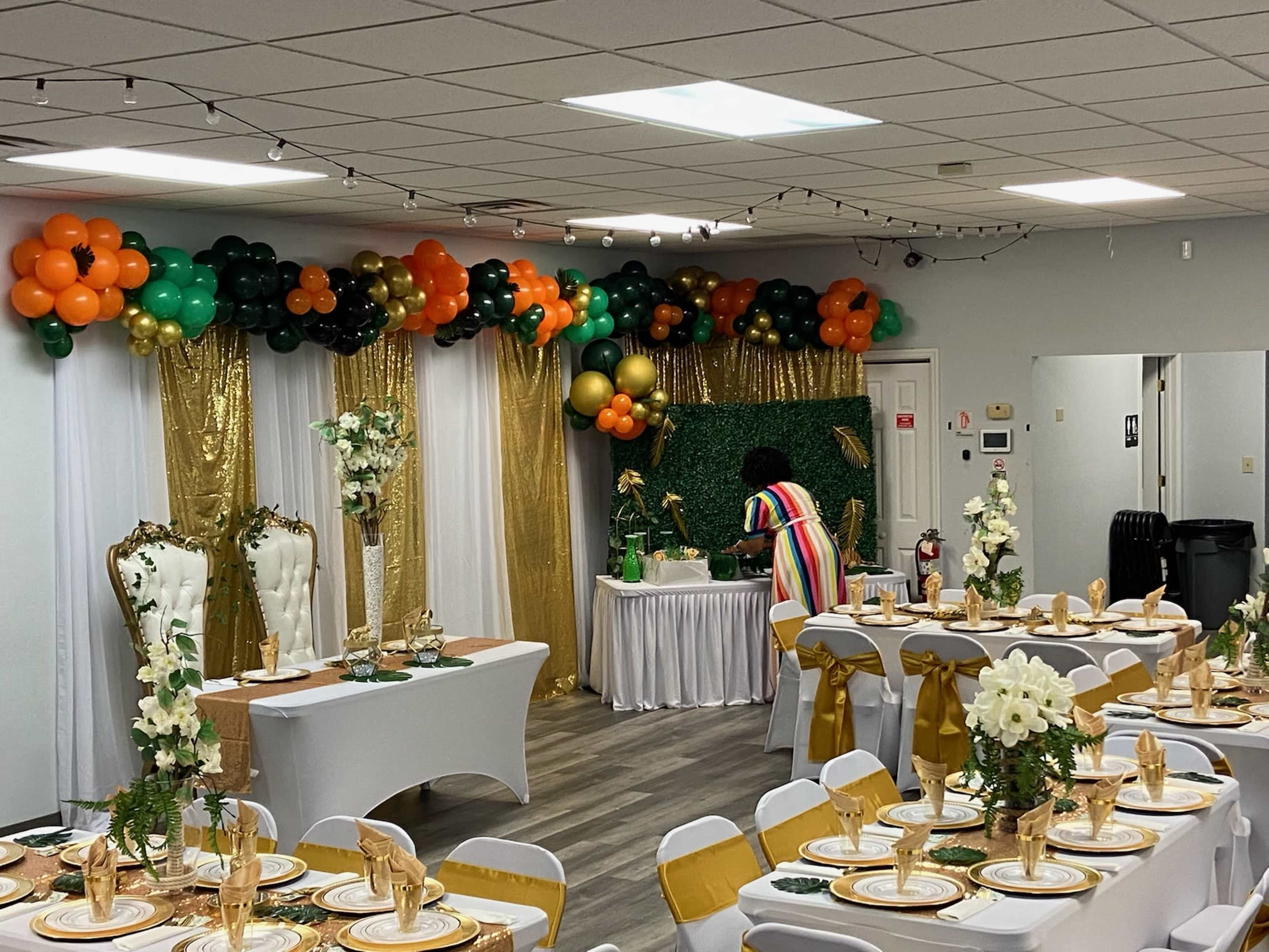A decorated event space with tables set for a formal gathering, featuring gold and green decorations, balloons, and a person arranging items on a table in the background.