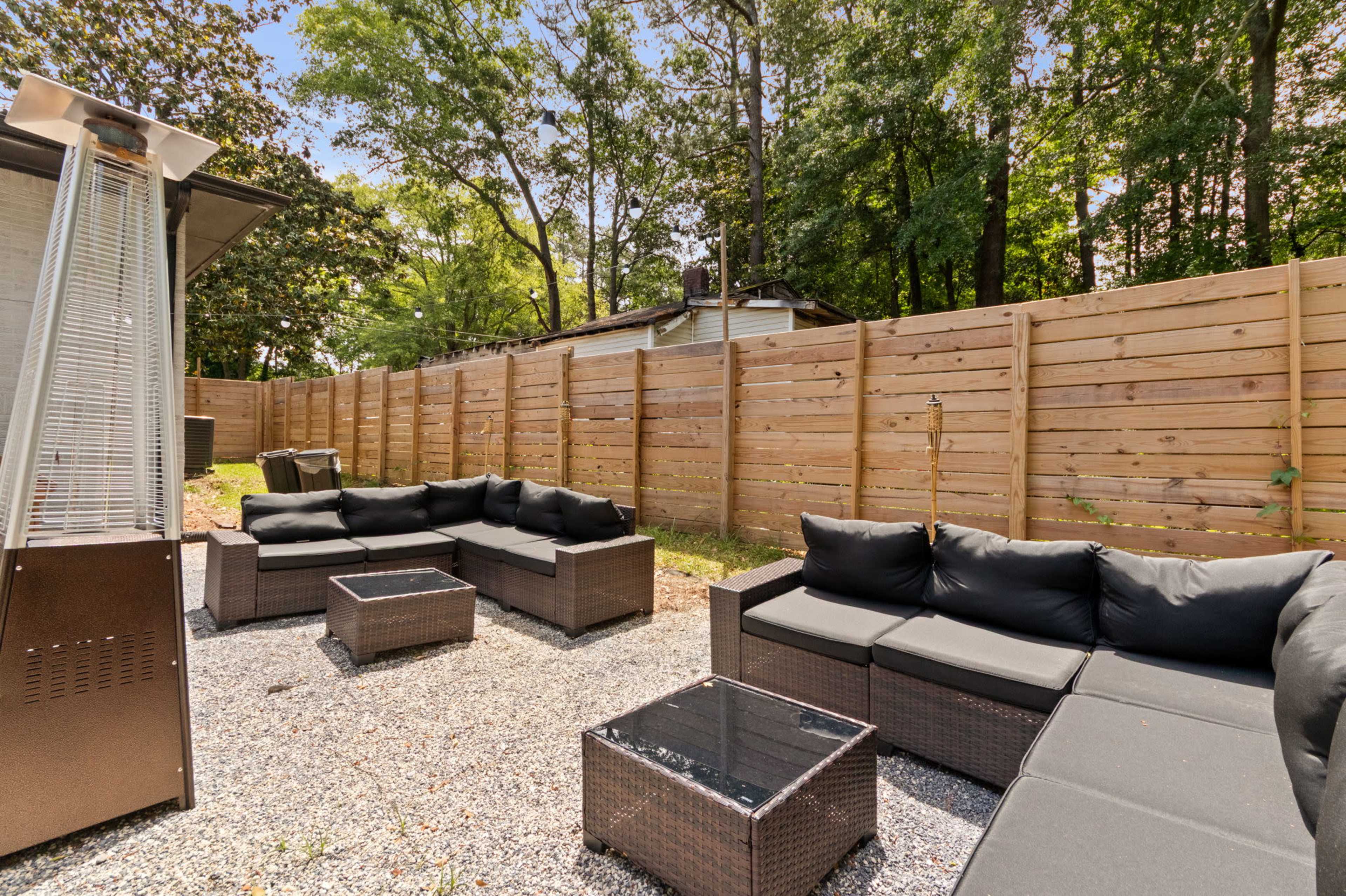 The image shows a cozy outdoor seating arrangement with black wicker furniture, a glass-top table, and a patio heater, surrounded by a wooden fence and lush trees.