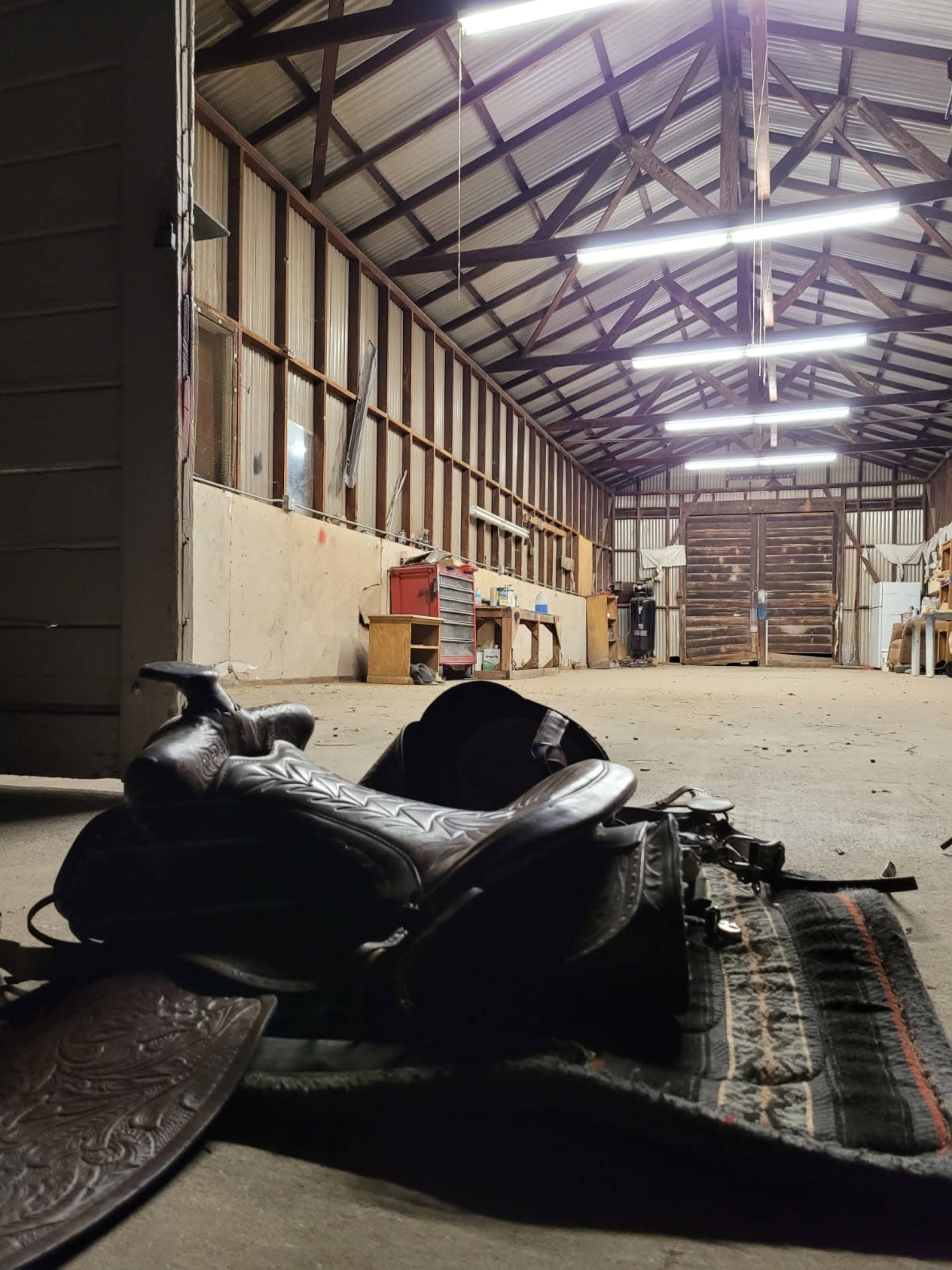 A saddle resting on a patterned rug inside a spacious, well-lit barn with wooden beams and tools lining the walls.