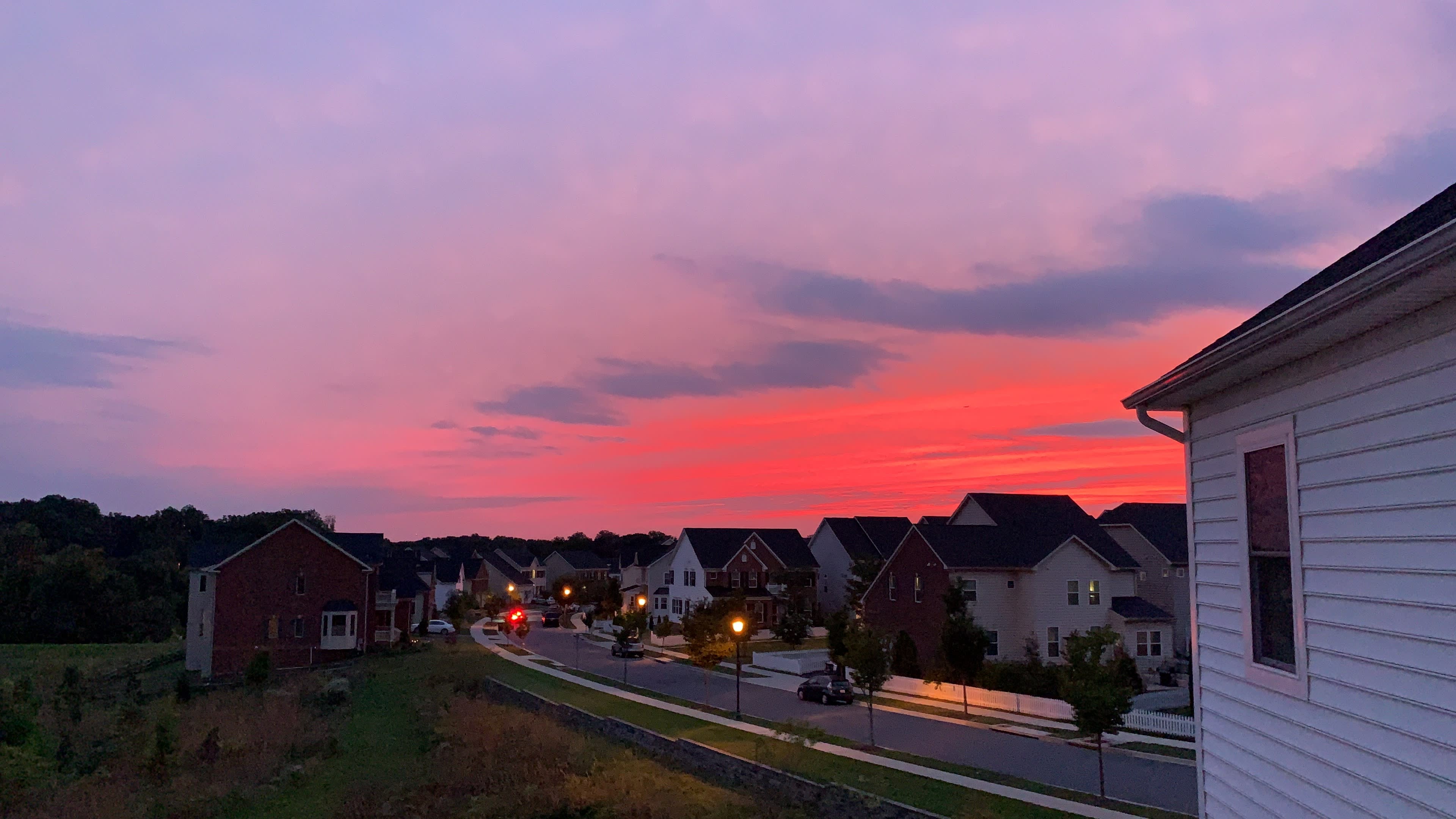 The scene shows a suburban neighborhood at dusk with houses lining a street under a colorful sunset.