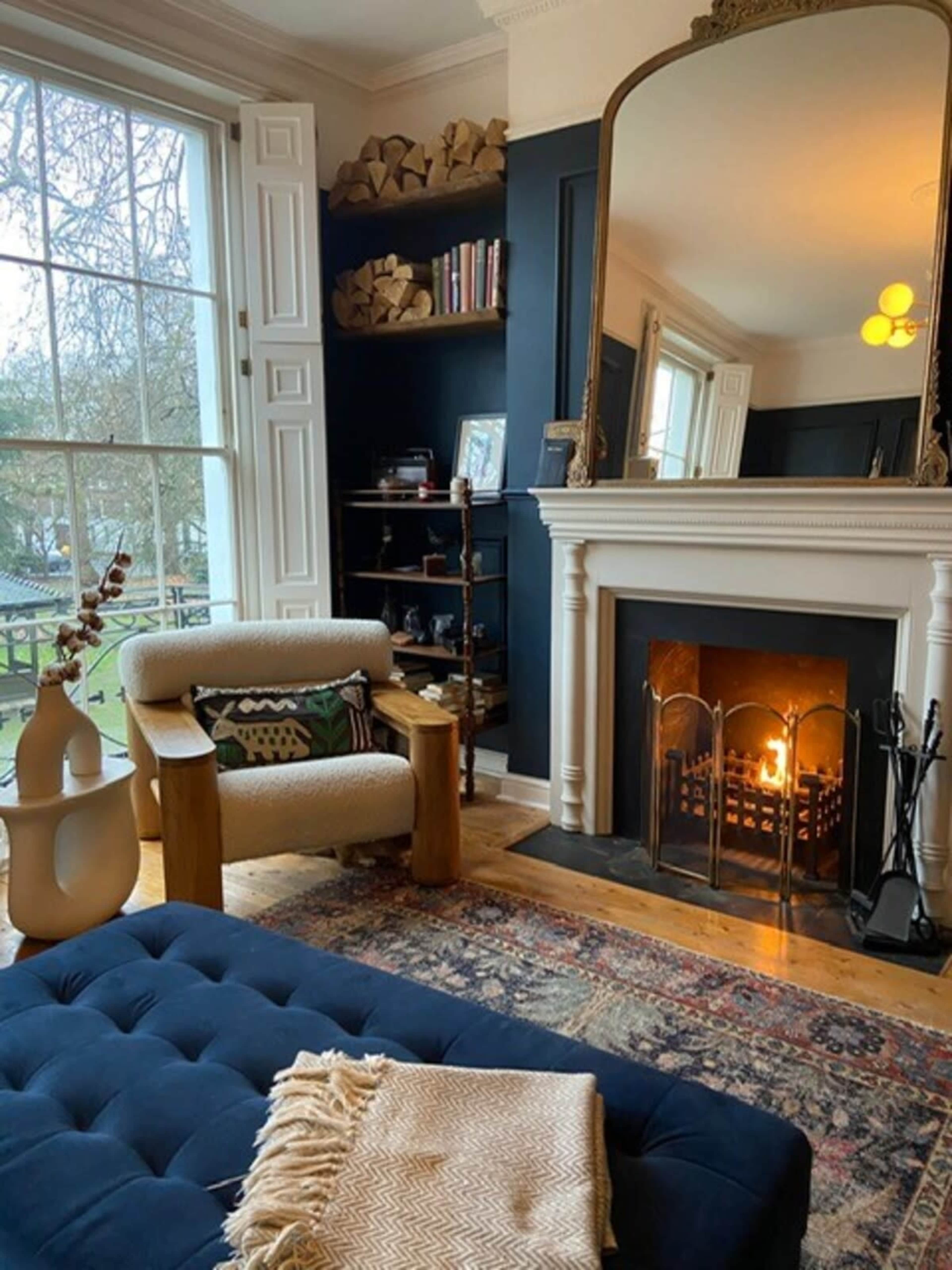 A cozy living room with a fireplace, a tufted blue ottoman, and a mirror above the mantel, overlooking a view outside through a large window.