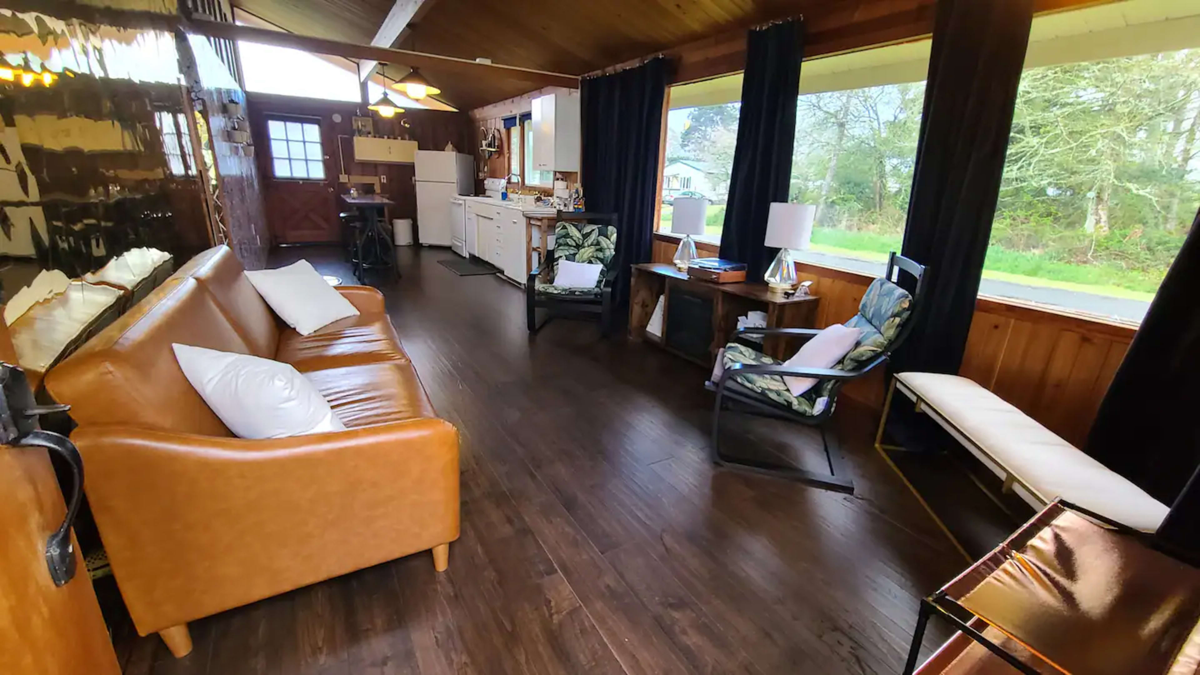 The interior of a cozy living space features a brown leather couch, wooden flooring, and large windows that provide a view of the outdoors.