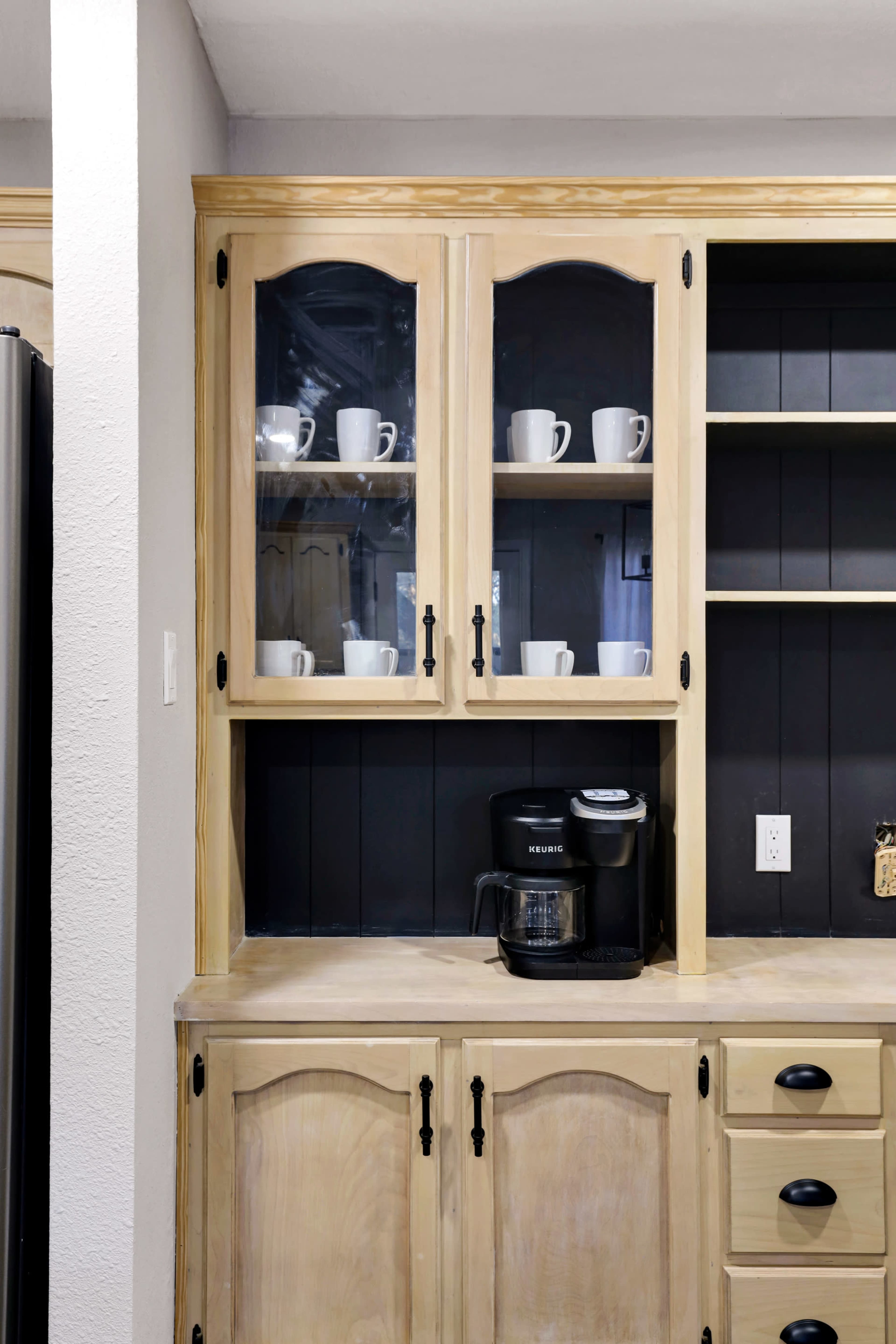 A wooden kitchen cabinet with glass doors displays white mugs, while a coffee maker is positioned on the countertop below.