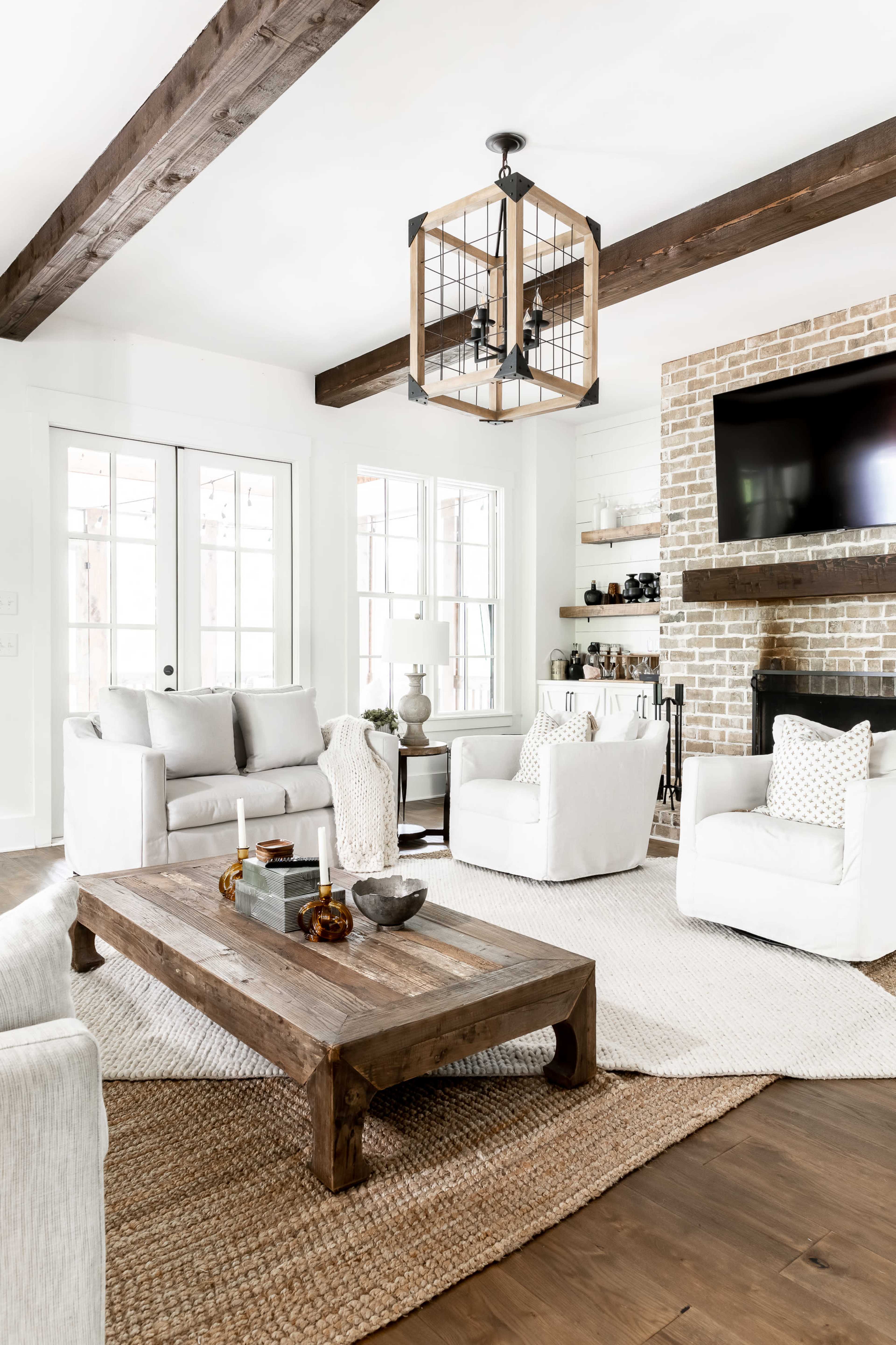 A modern living room features a brick fireplace, a wooden coffee table, and white upholstered furniture arranged around a woven area rug.