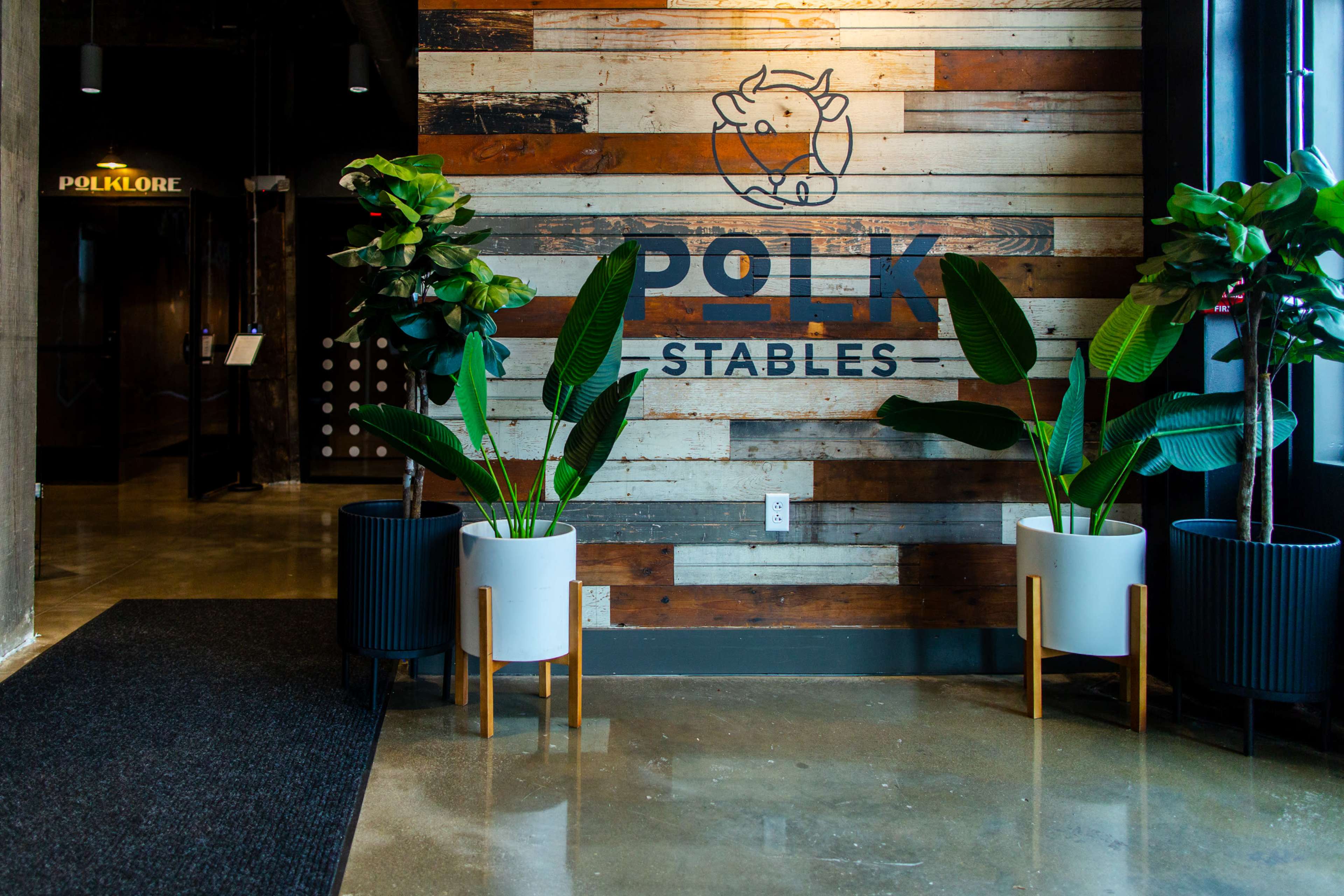 The image shows a modern lobby area featuring a wooden accent wall with the words "Polk Stables," two large potted plants, and a shiny concrete floor.