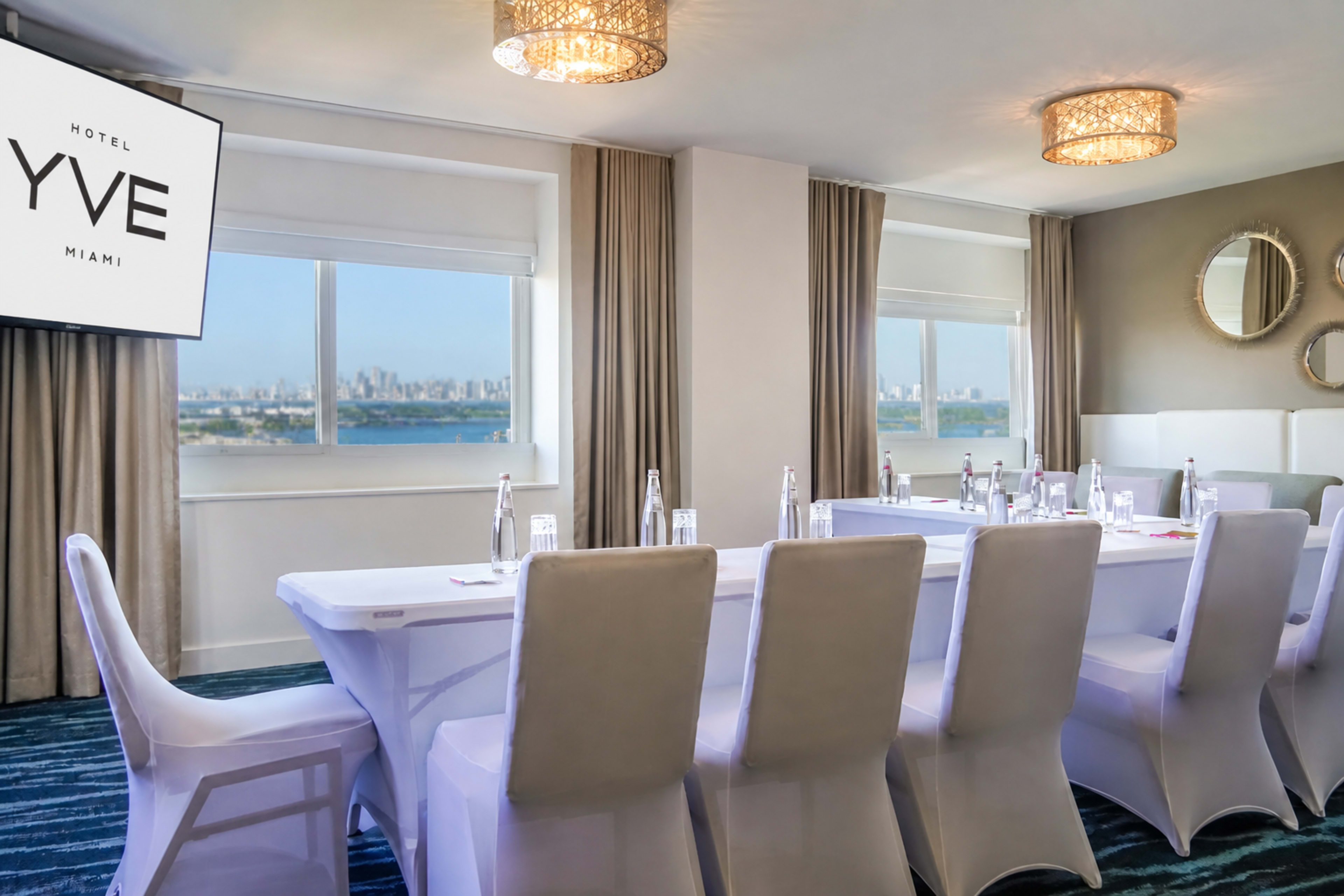 The image shows a modern conference room with a long table set for a meeting, featuring a scenic view of a city skyline through large windows.
