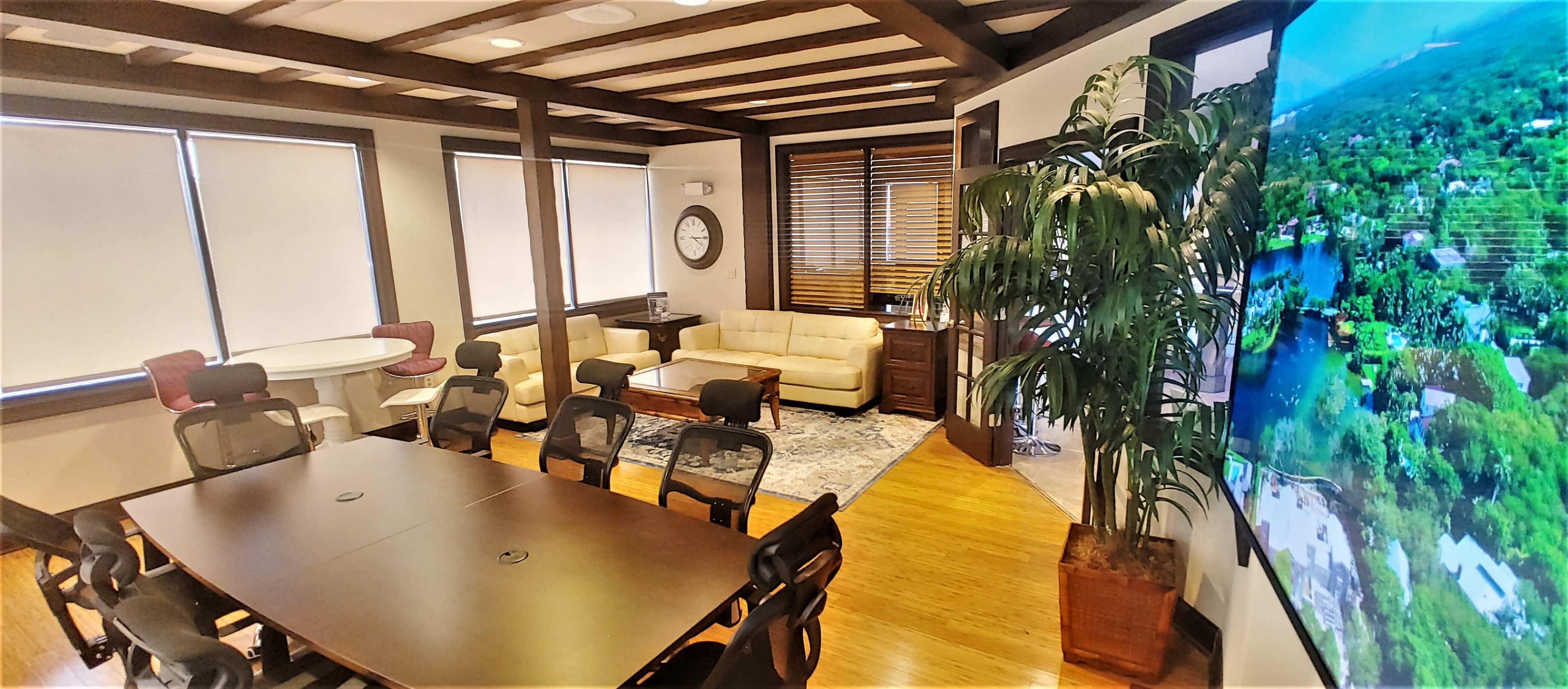 The image shows a modern office space featuring a conference table with chairs, a lounge area with a sofa, and large windows with blinds, along with a wall-mounted display screen.