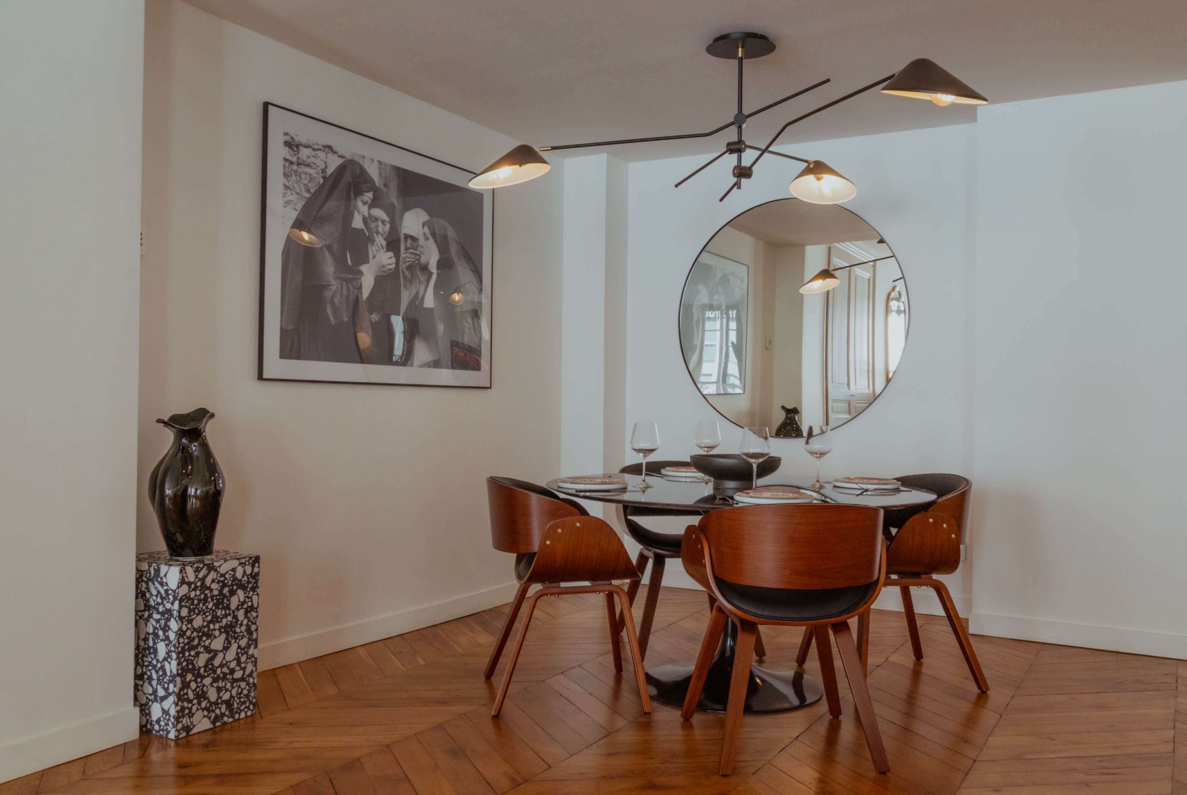 A modern dining area features a round table with four wooden chairs, surrounded by a decorative vase and a large circular mirror on the wall.