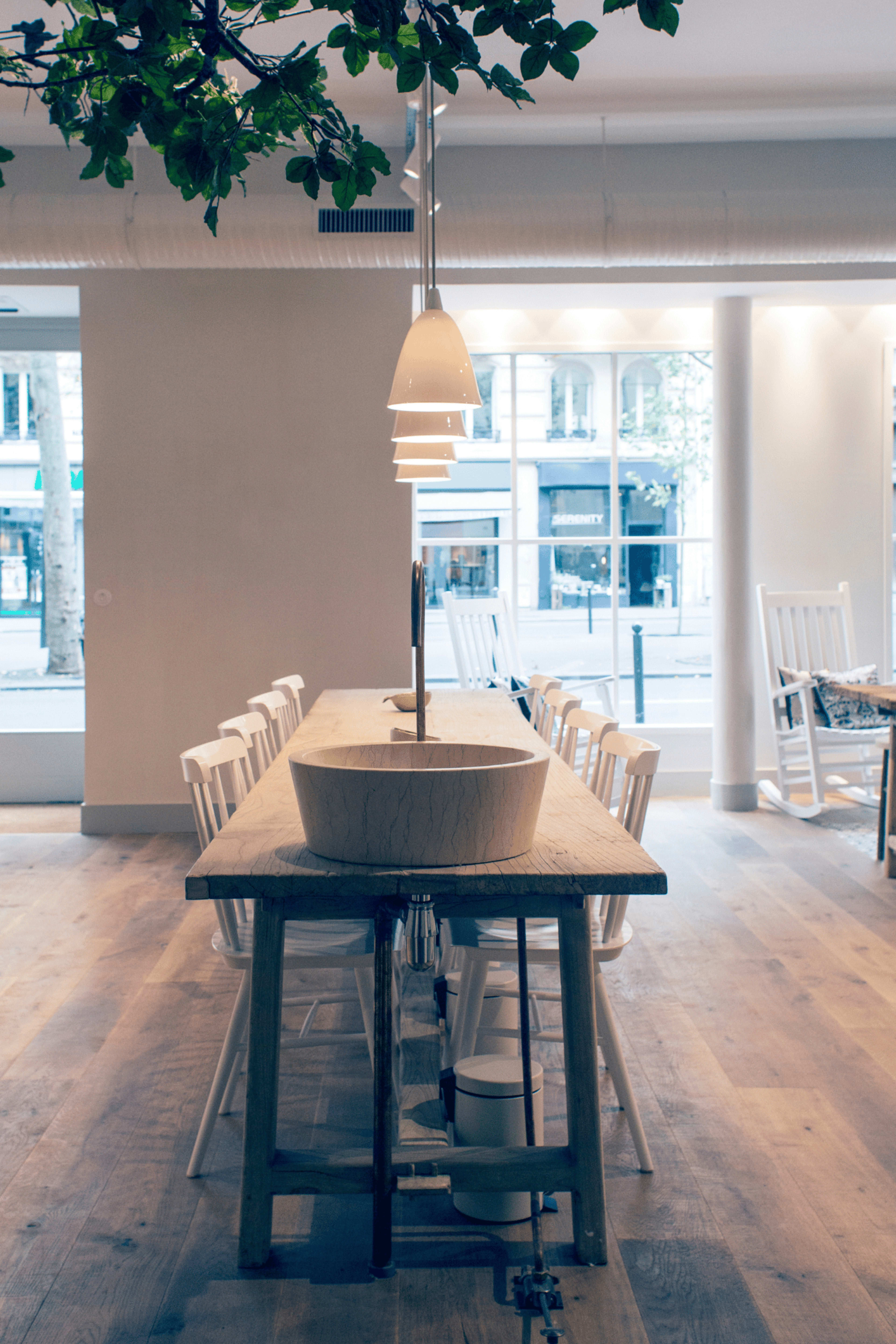 A wooden dining table with a central bowl is surrounded by white chairs in a bright, modern space with large windows.