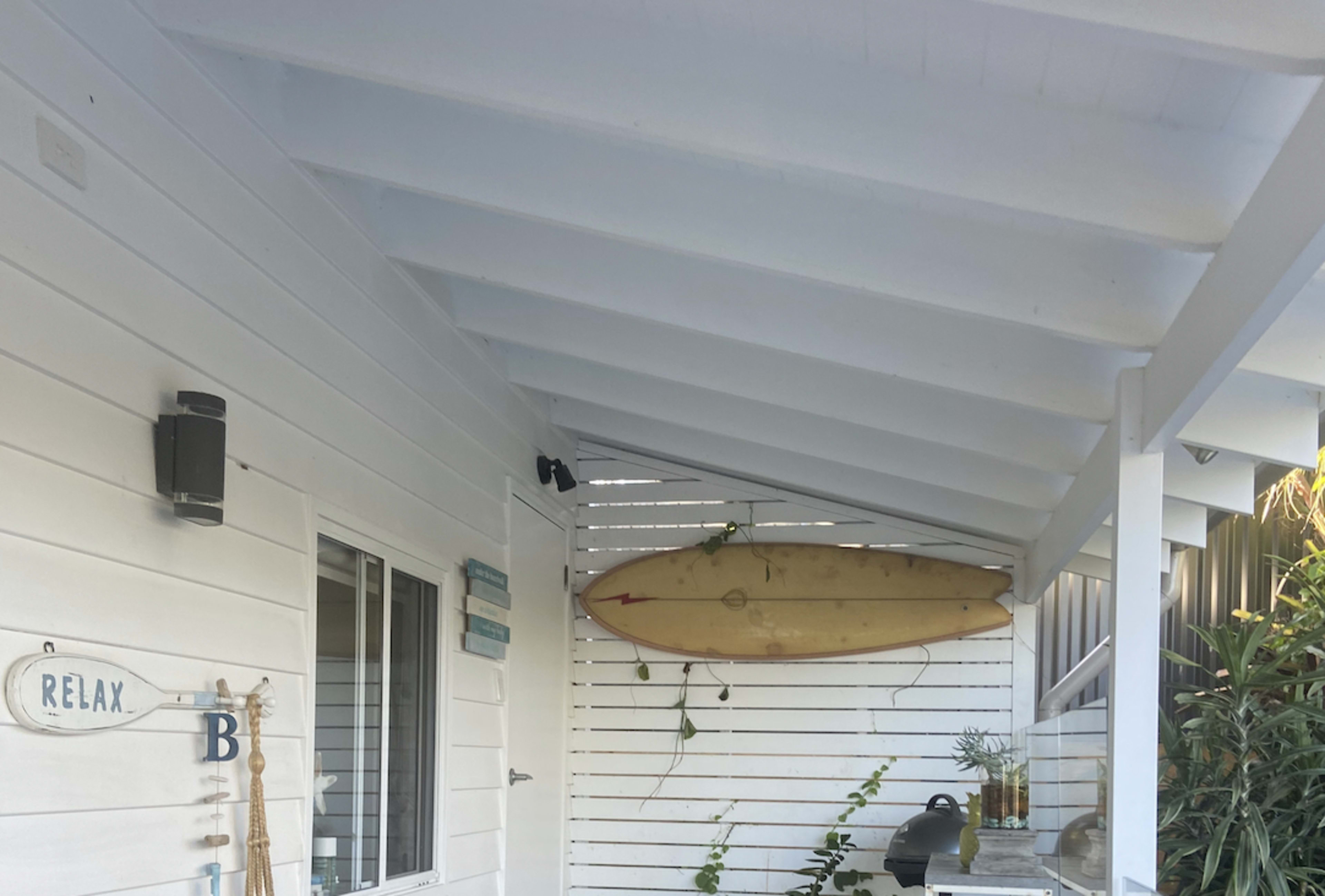 A porch with white walls, a surfboard mounted on the ceiling, and a grill on the right.