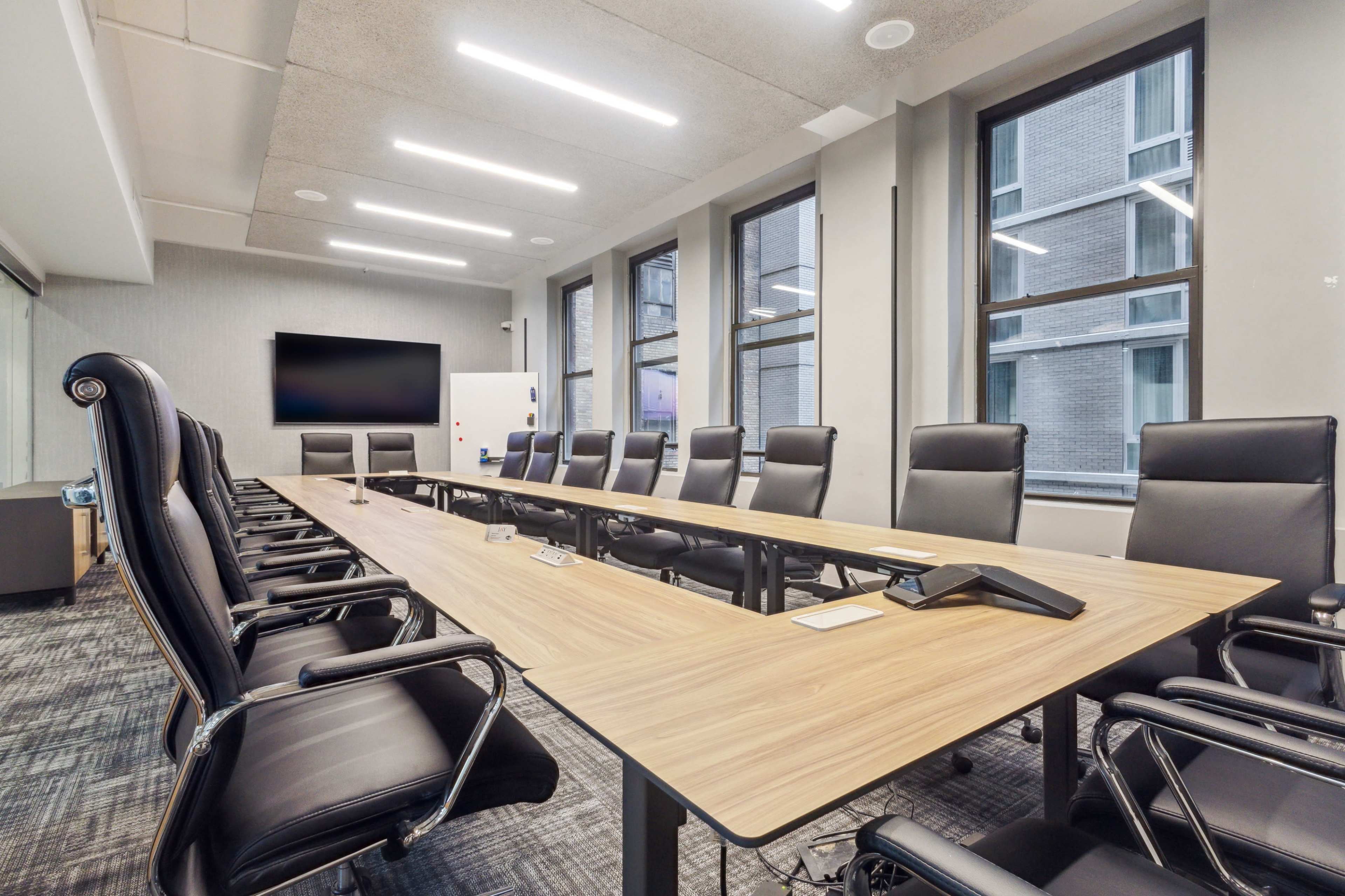 A modern conference room features a long wooden table surrounded by ergonomic black chairs and large windows providing natural light.