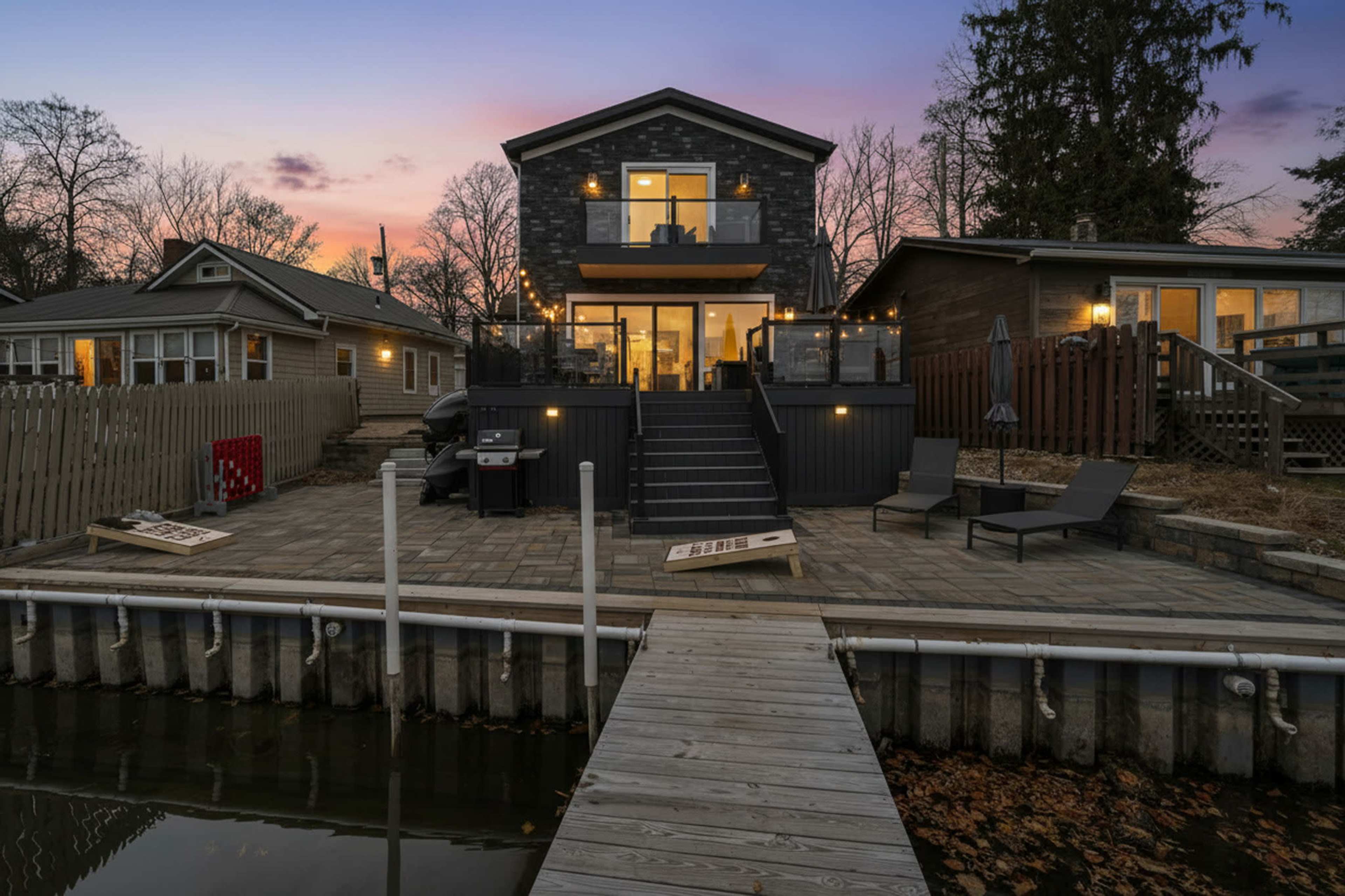 The image shows a modern waterfront house with a large deck and outdoor seating area, situated beside a calm body of water at dusk.