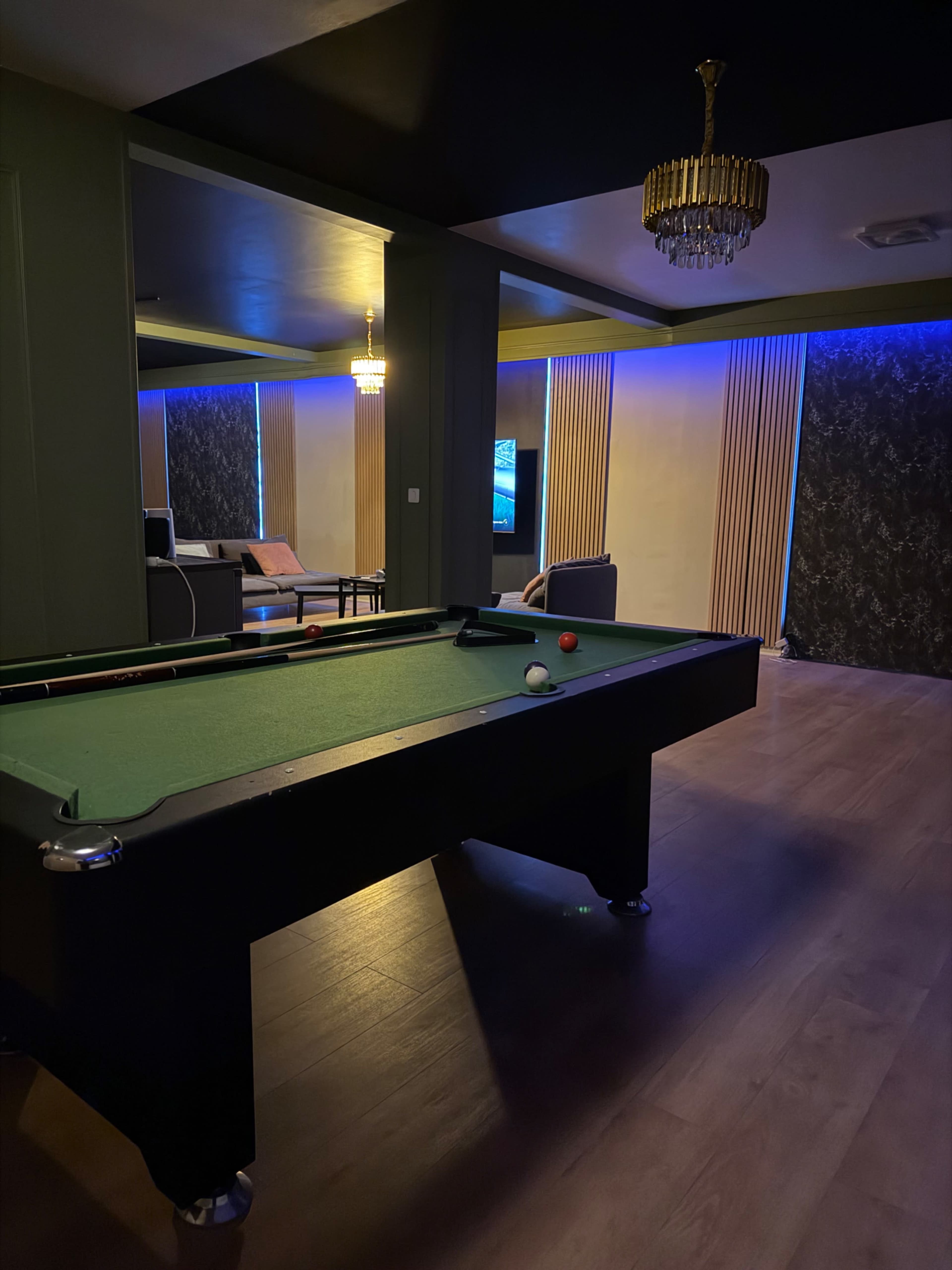 The image shows a billiards table in a dimly lit room with modern decor, featuring a chandelier and ambient lighting along the walls.