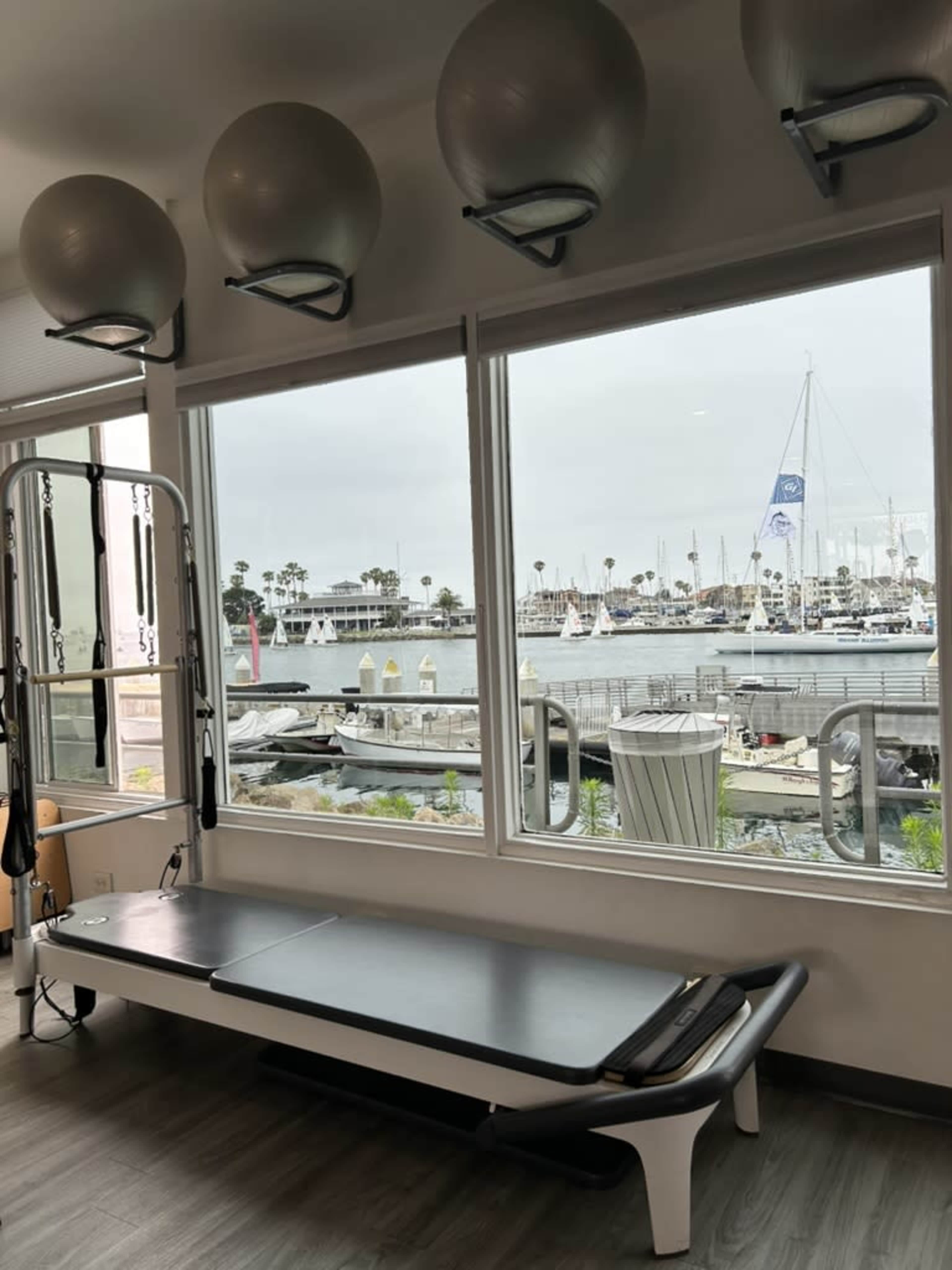 The image shows a gym studio with a reformer machine in front of large windows overlooking a marina with boats and a cloudy sky.