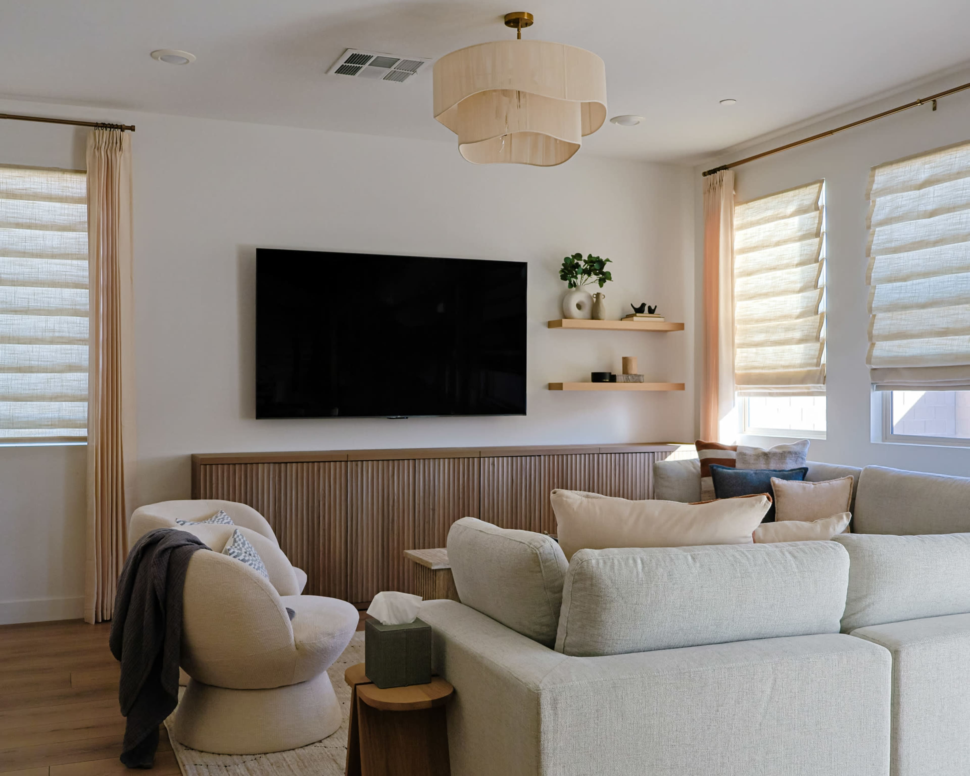 The image shows a modern living room featuring a large flat-screen TV mounted on the wall, a sectional sofa, and light window treatments, with wooden shelves displaying decorative items.