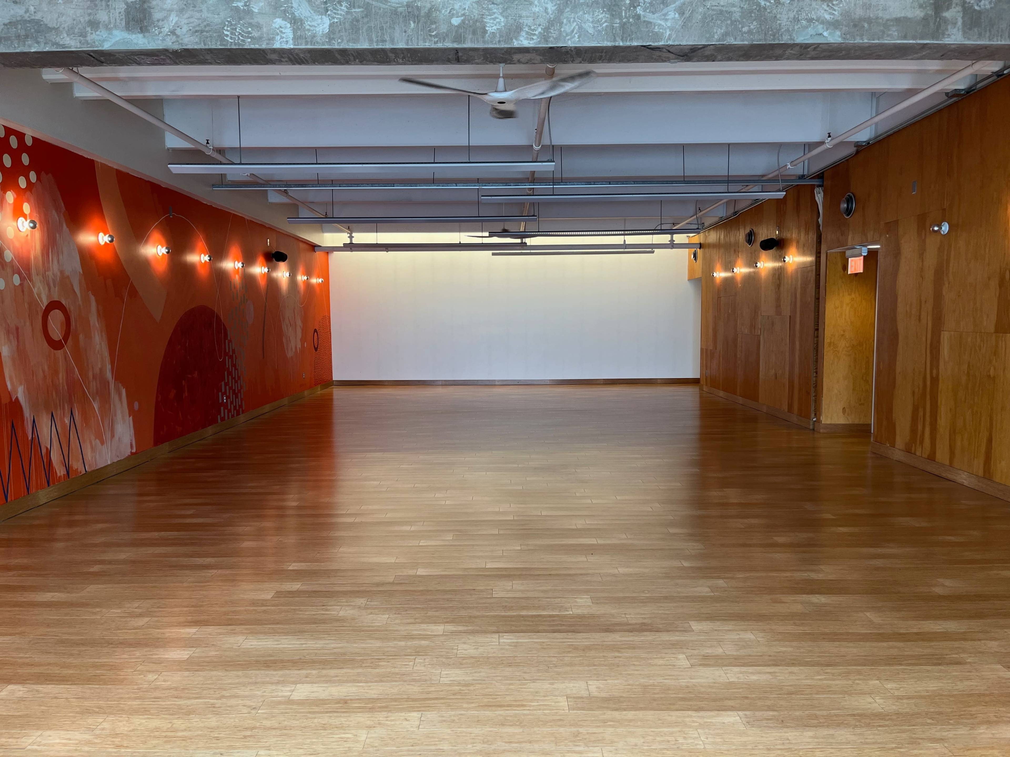 A spacious room with wooden flooring, adorned with a vibrant red and white mural on one wall and a bright, illuminated area at the far end.