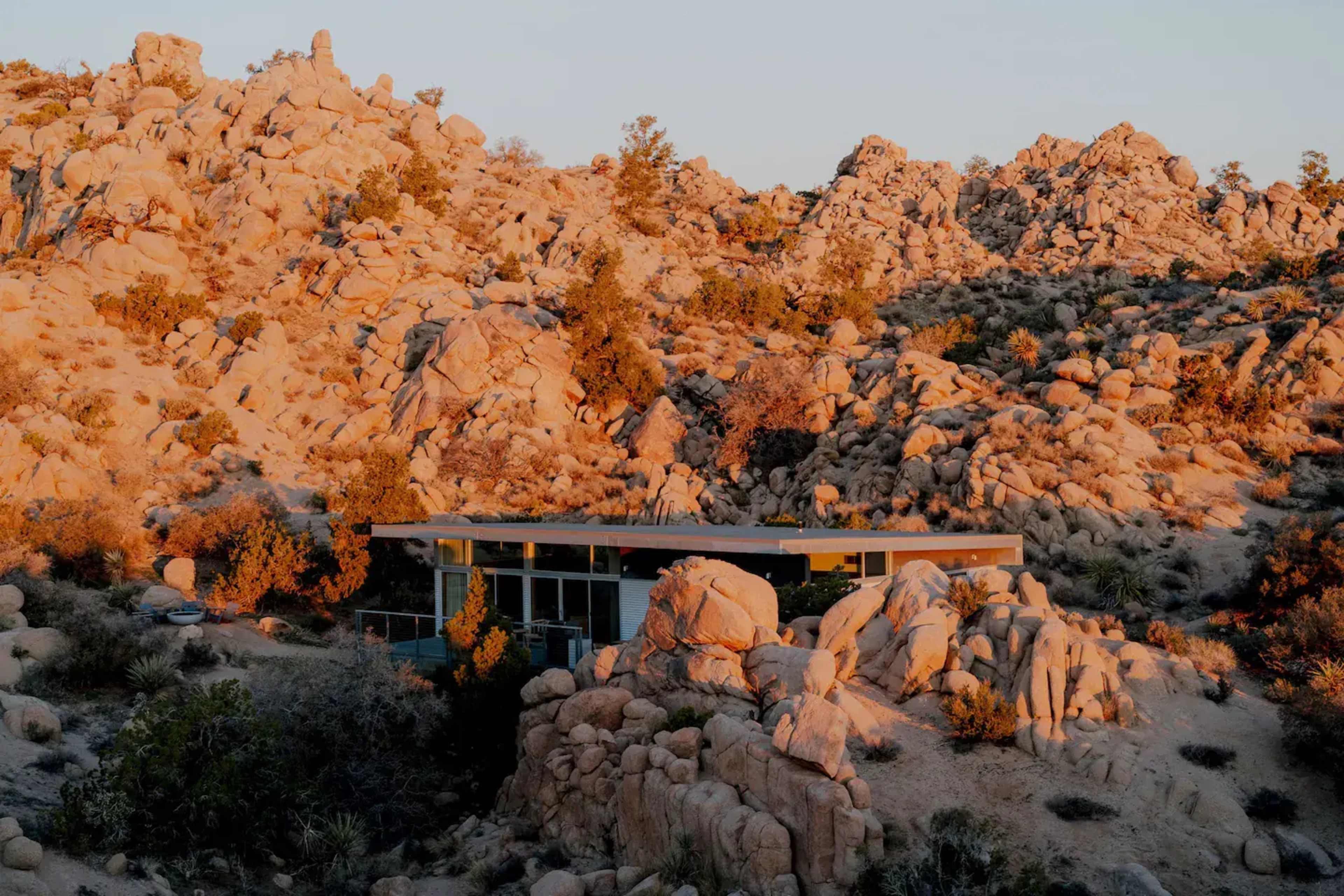 A modern house is nestled among large boulders and desert vegetation in a rocky landscape.