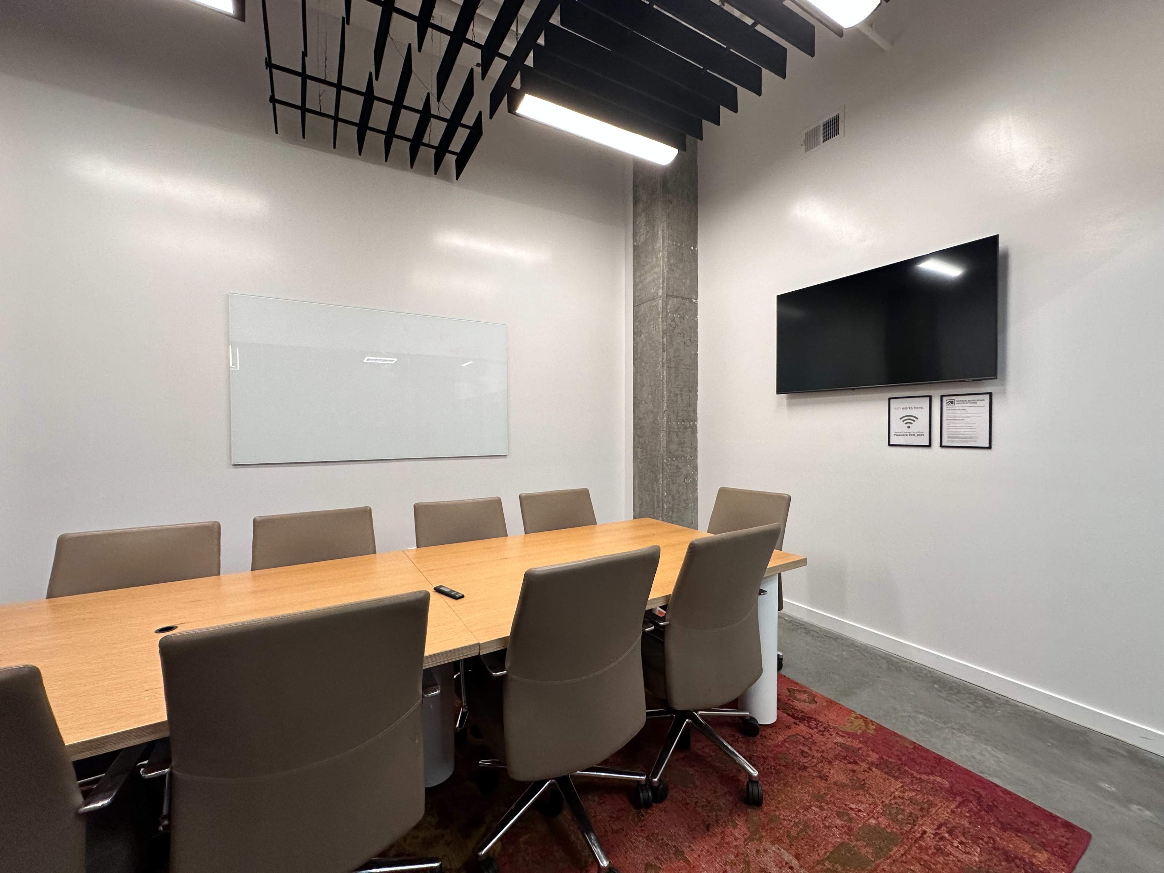 A conference room features a long wooden table surrounded by eight chairs, with a large wall-mounted screen and a whiteboard on the adjacent wall.