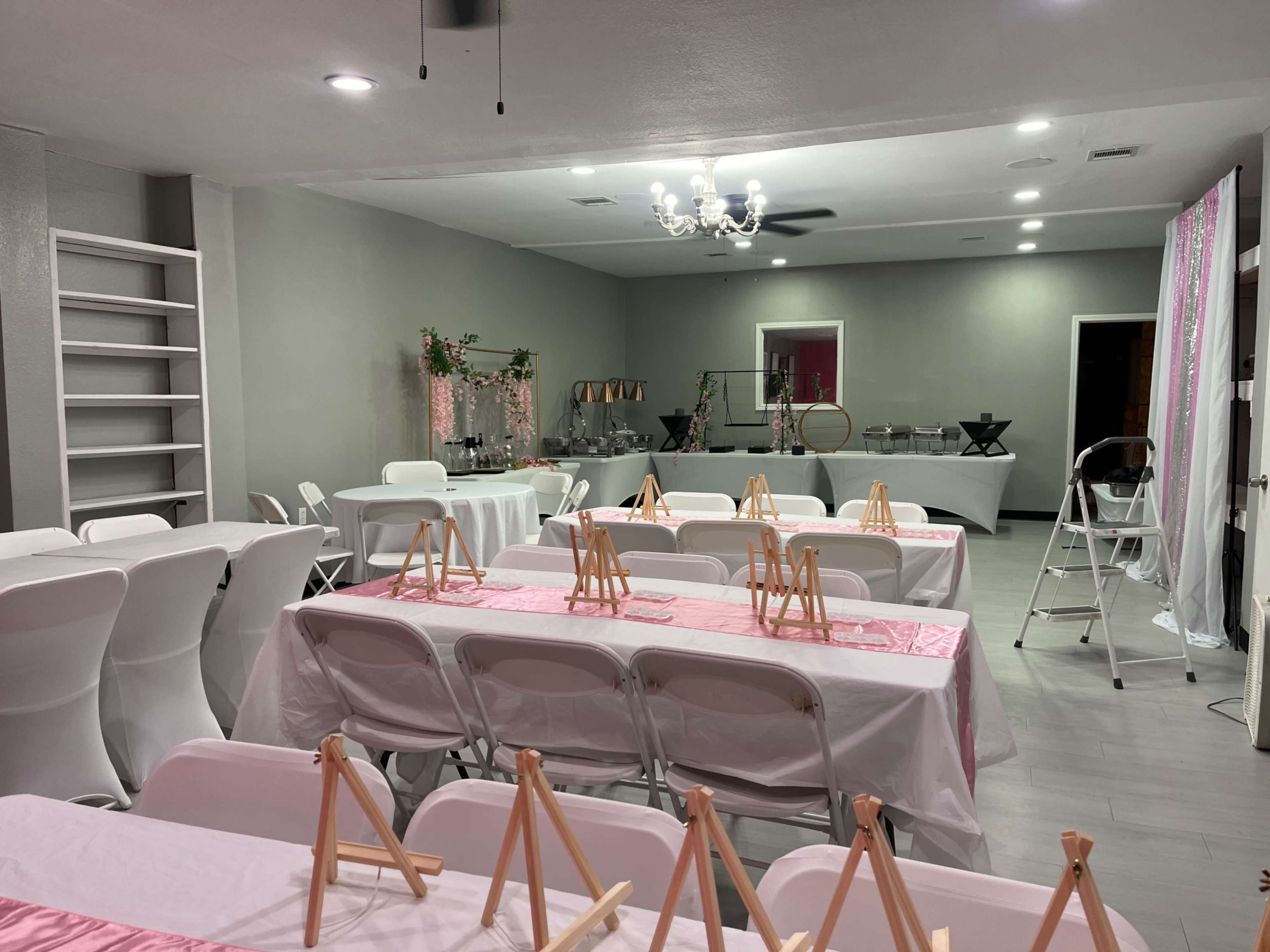 The image shows an event space set up with white tables and chairs, decorated with pink table runners and easels for a gathering or party.