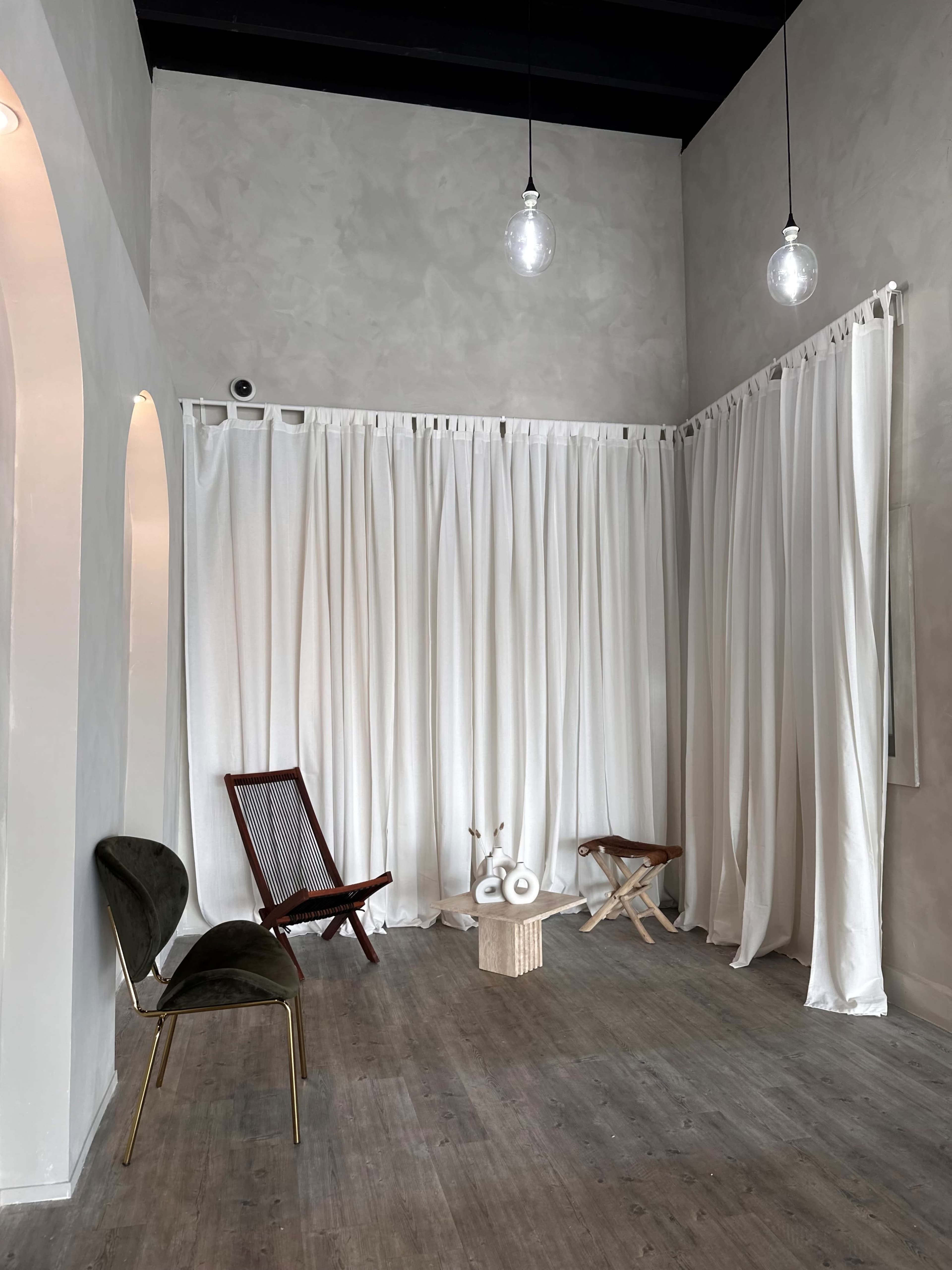 A corner of an interior room featuring white curtains, a wooden chair, a folding stool, and decorative objects on a small pedestal.