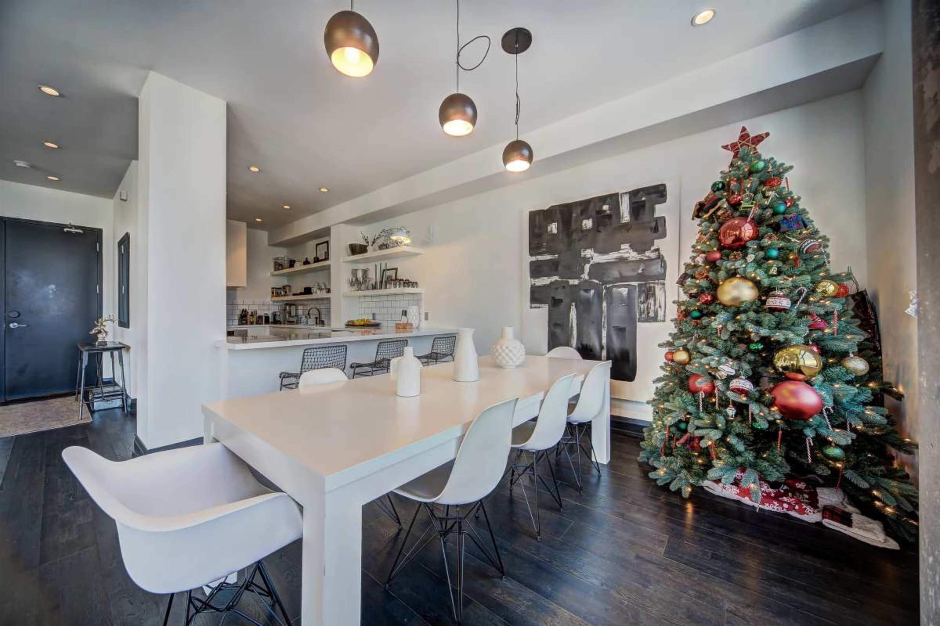 A modern dining area features a long white table surrounded by chairs, with a decorated Christmas tree in the corner and an open kitchen in the background.