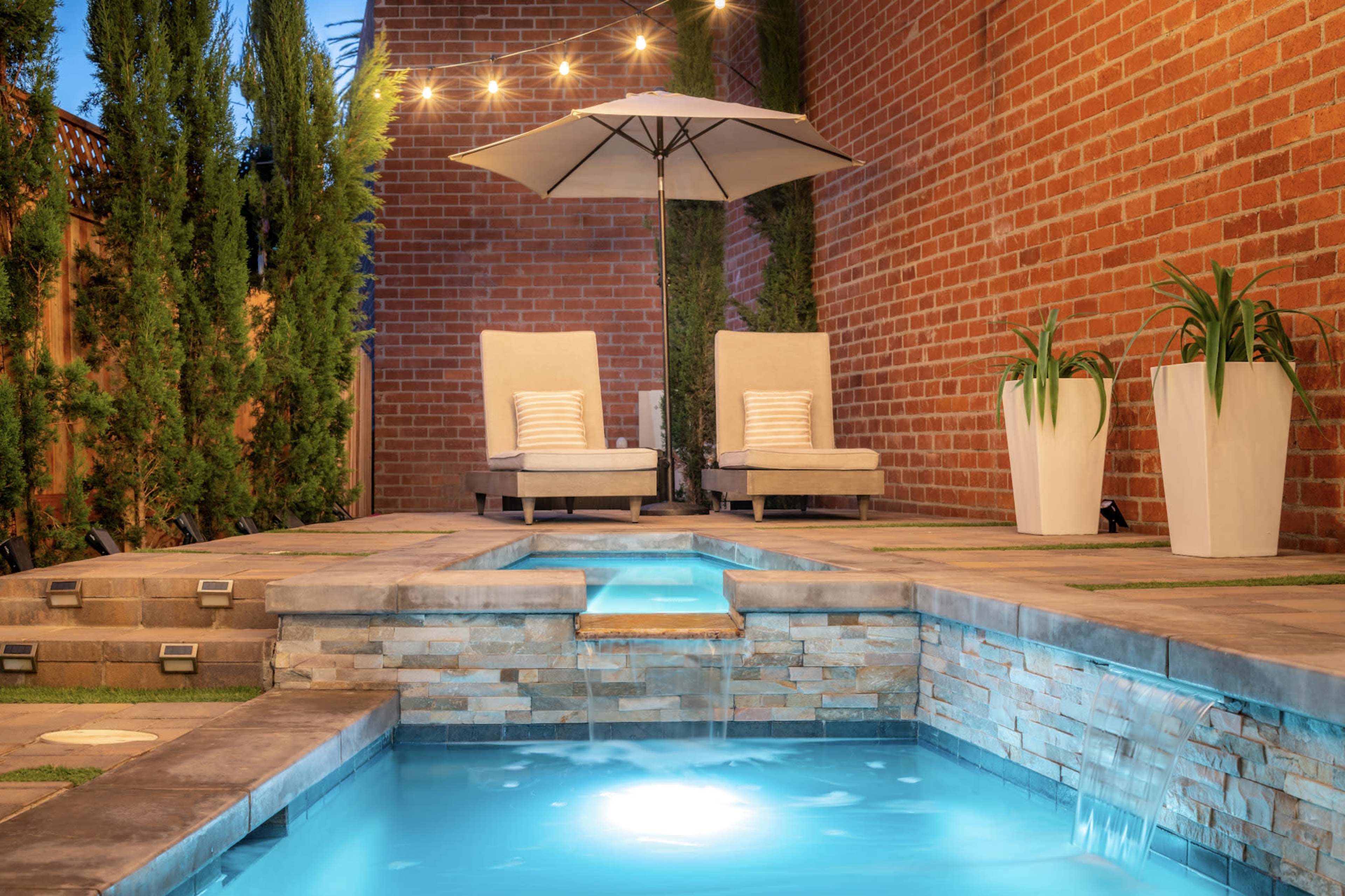 A small pool area features two lounge chairs under an umbrella, surrounded by brick walls and potted plants.