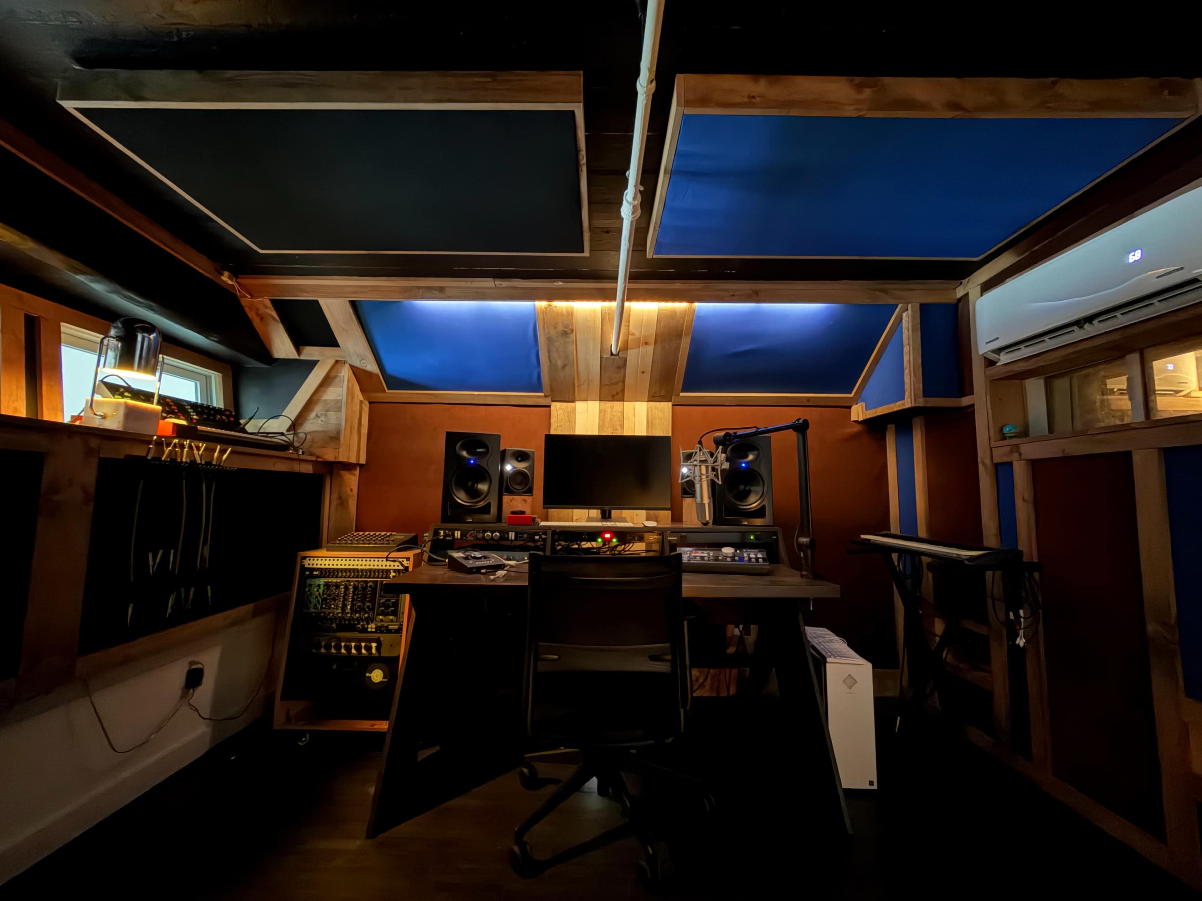The image shows a recording studio with wooden walls, a central desk equipped with audio equipment, and soundproofing panels on the ceiling.