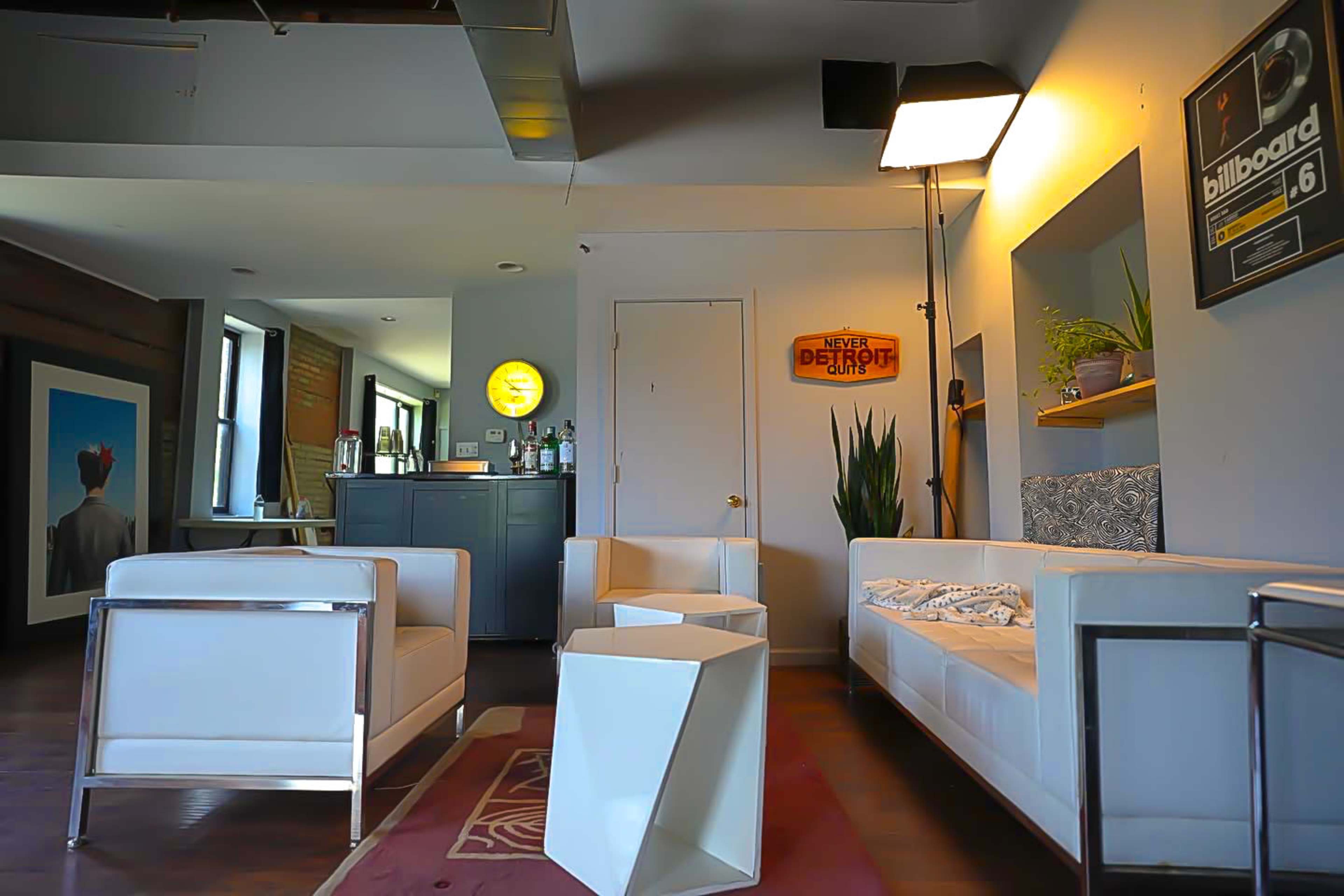 The image shows a modern lounge area featuring two white sofas, a geometric white side table, a bar counter with liquor bottles, and framed artwork on the walls.
