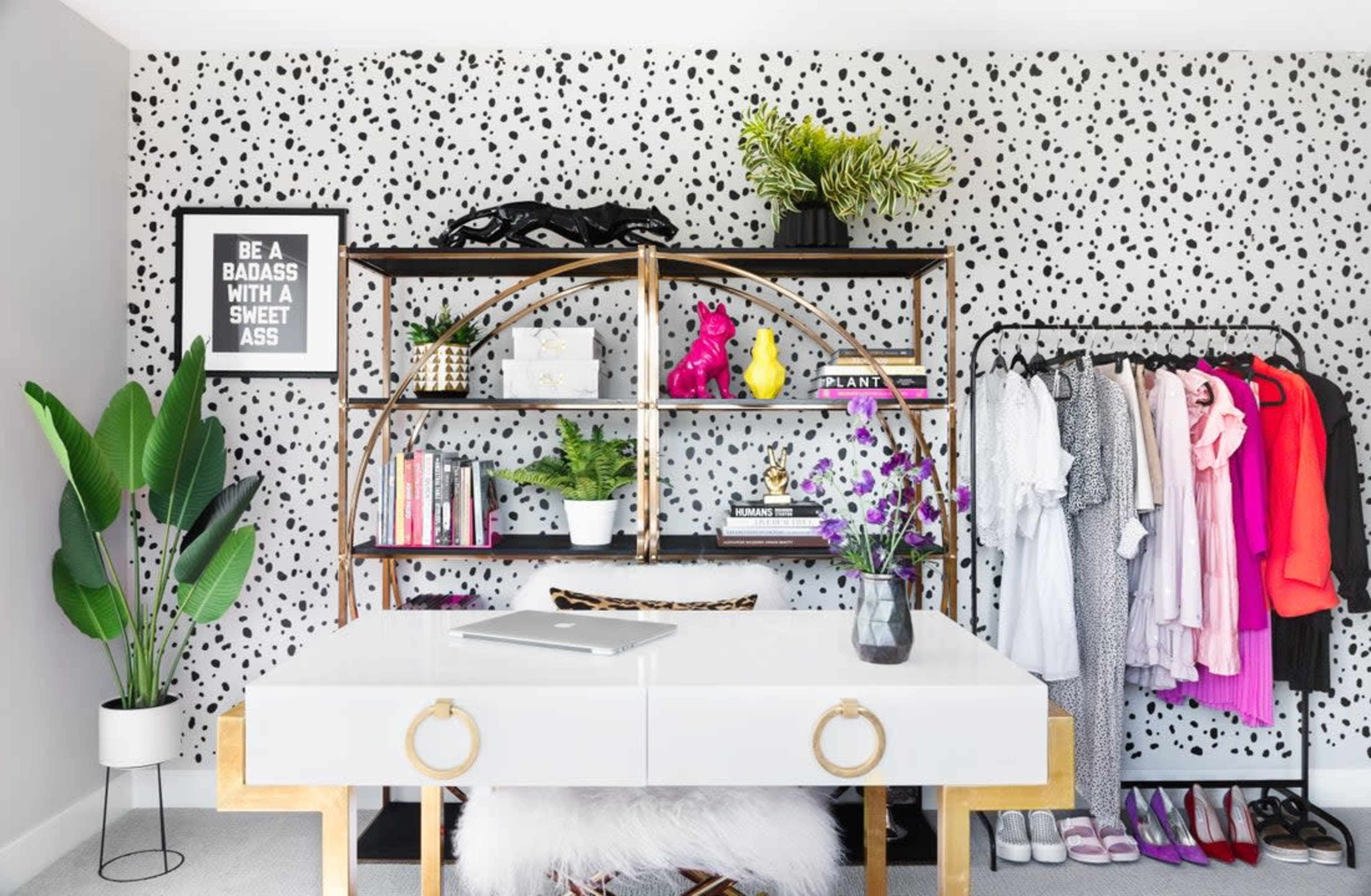 The image shows a modern workspace featuring a white desk with gold accents, a stylish bookshelf filled with books and decorative items, a clothing rack with various garments, and potted plants against a polka-dotted wall.