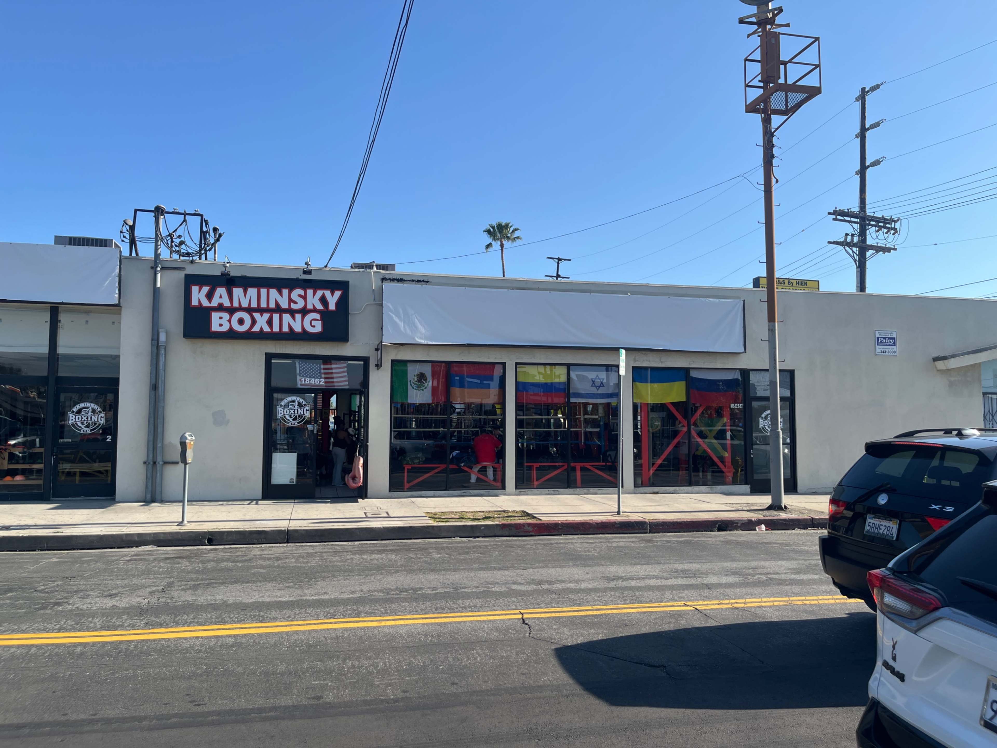 The image shows the exterior of a boxing gym named Kaminsky Boxing, featuring a row of international flags displayed in the front windows.