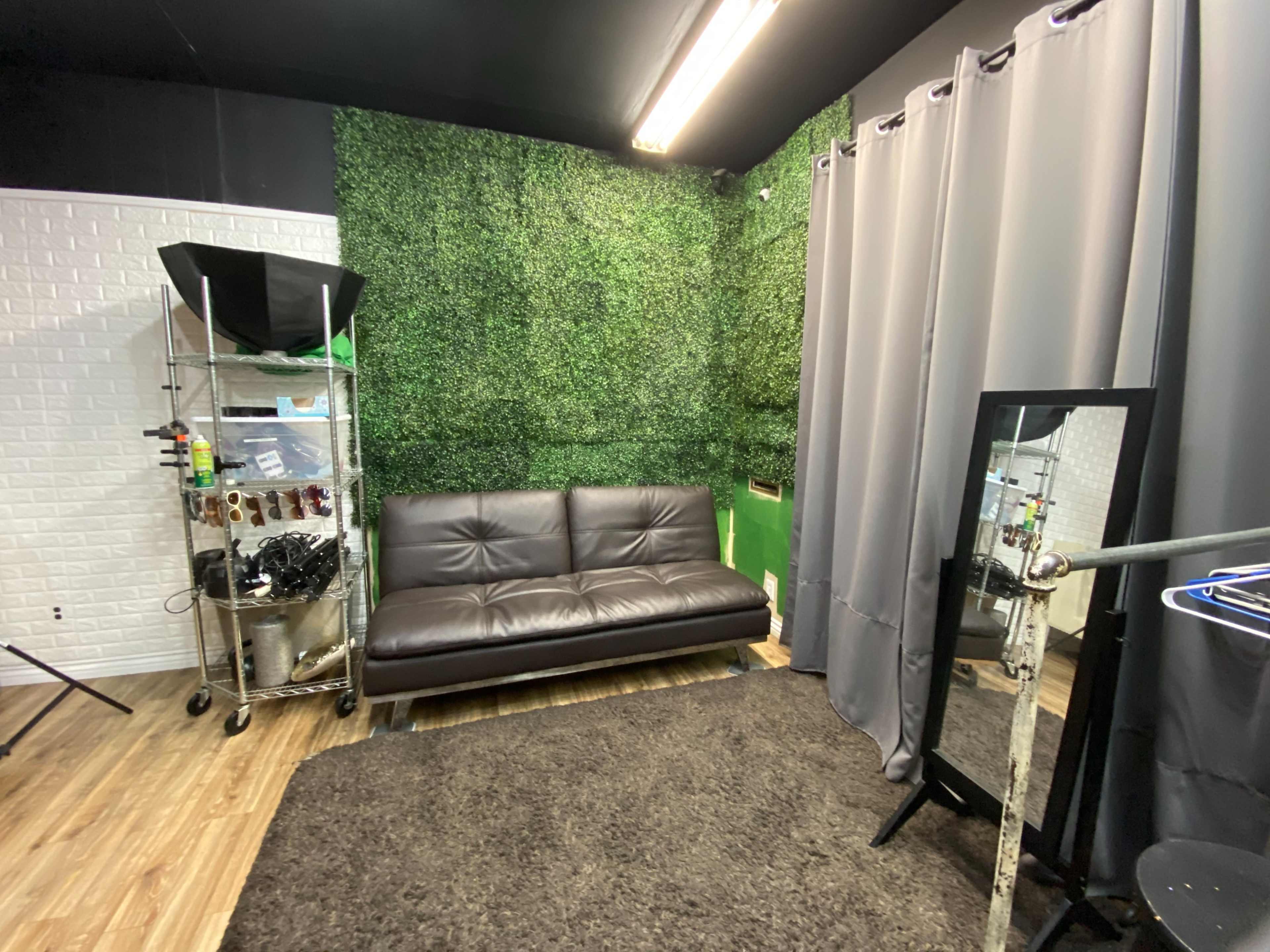 A small studio space featuring a brown sofa, a green hedge accent wall, gray curtains, and a mirror.