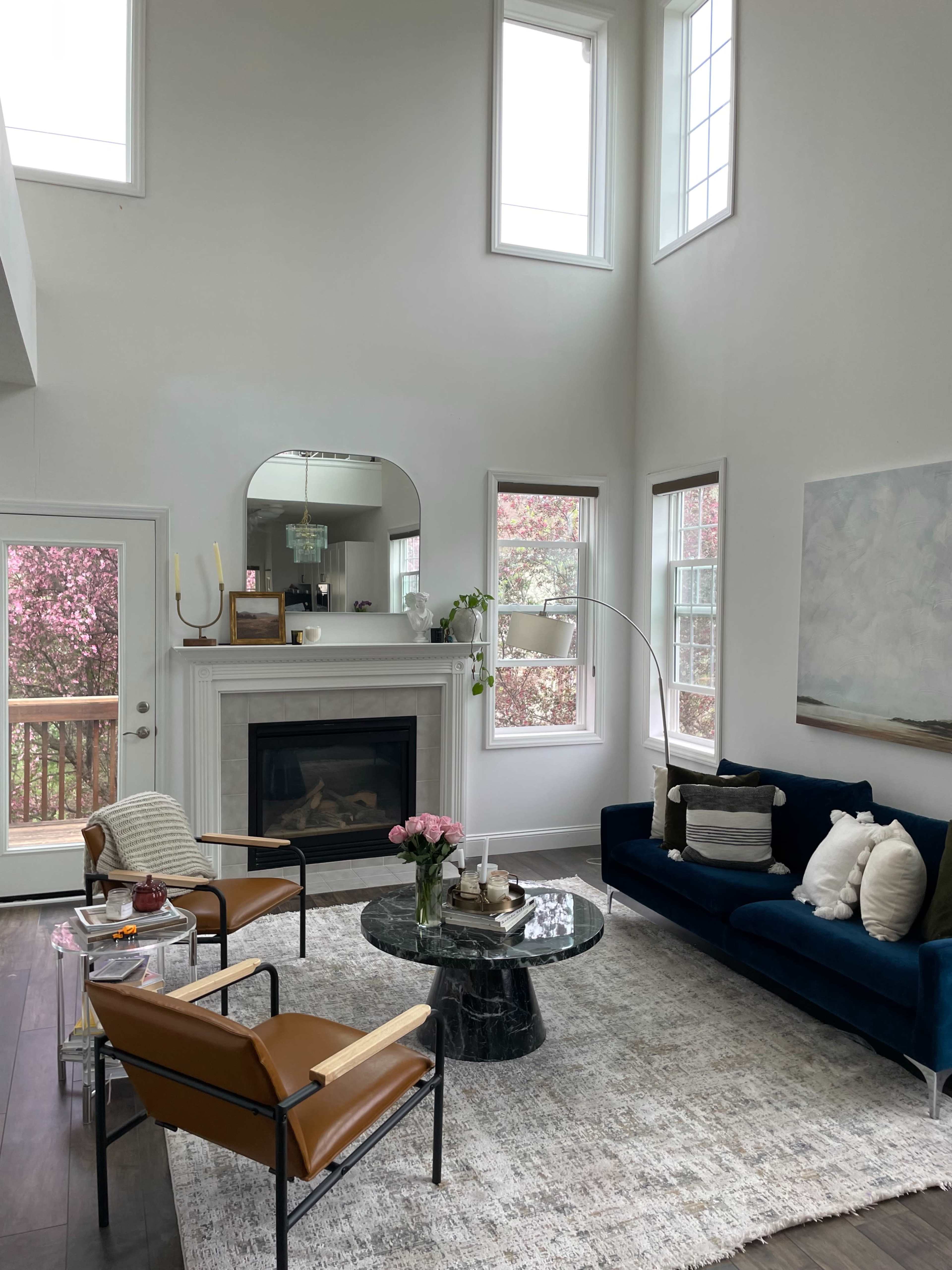A modern living room features a navy blue sofa, two brown armchairs, a round black marble coffee table, and a fireplace, with large windows allowing natural light and a view of blooming trees outside.
