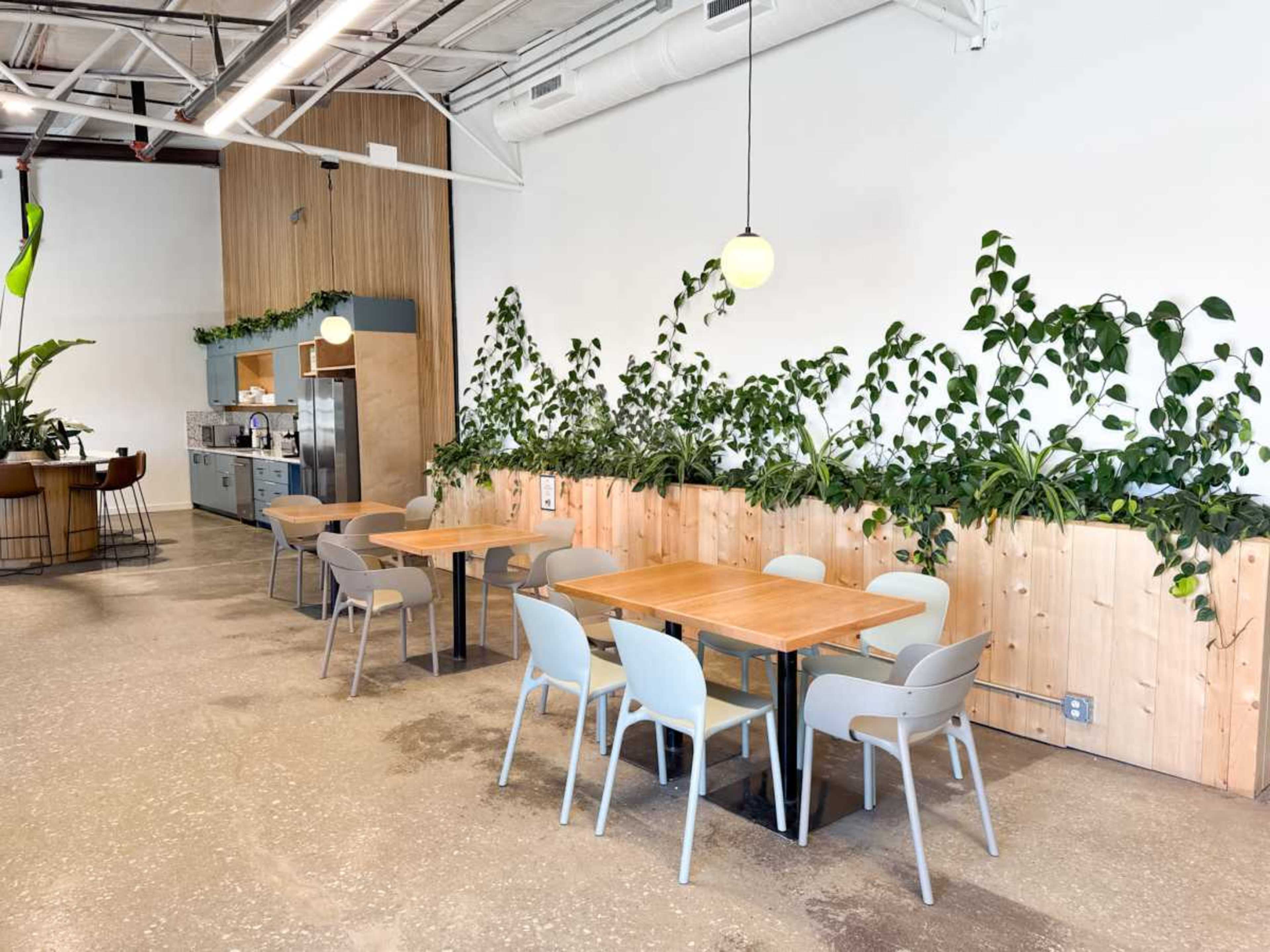 The image shows a modern dining area with wooden tables and gray chairs, surrounded by greenery and a kitchenette in the background.